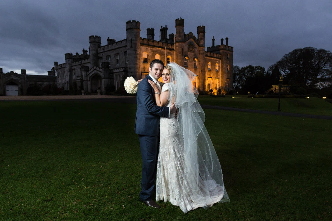 newlyweds embrace at dusk in front of the castle on the lawn with the castle lit up in the background