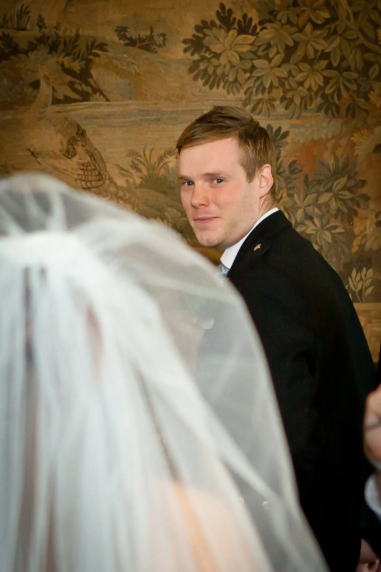 Groom in a black suit glancing back over his shoulder at the bride, whose white veil is softly focused in the foreground.