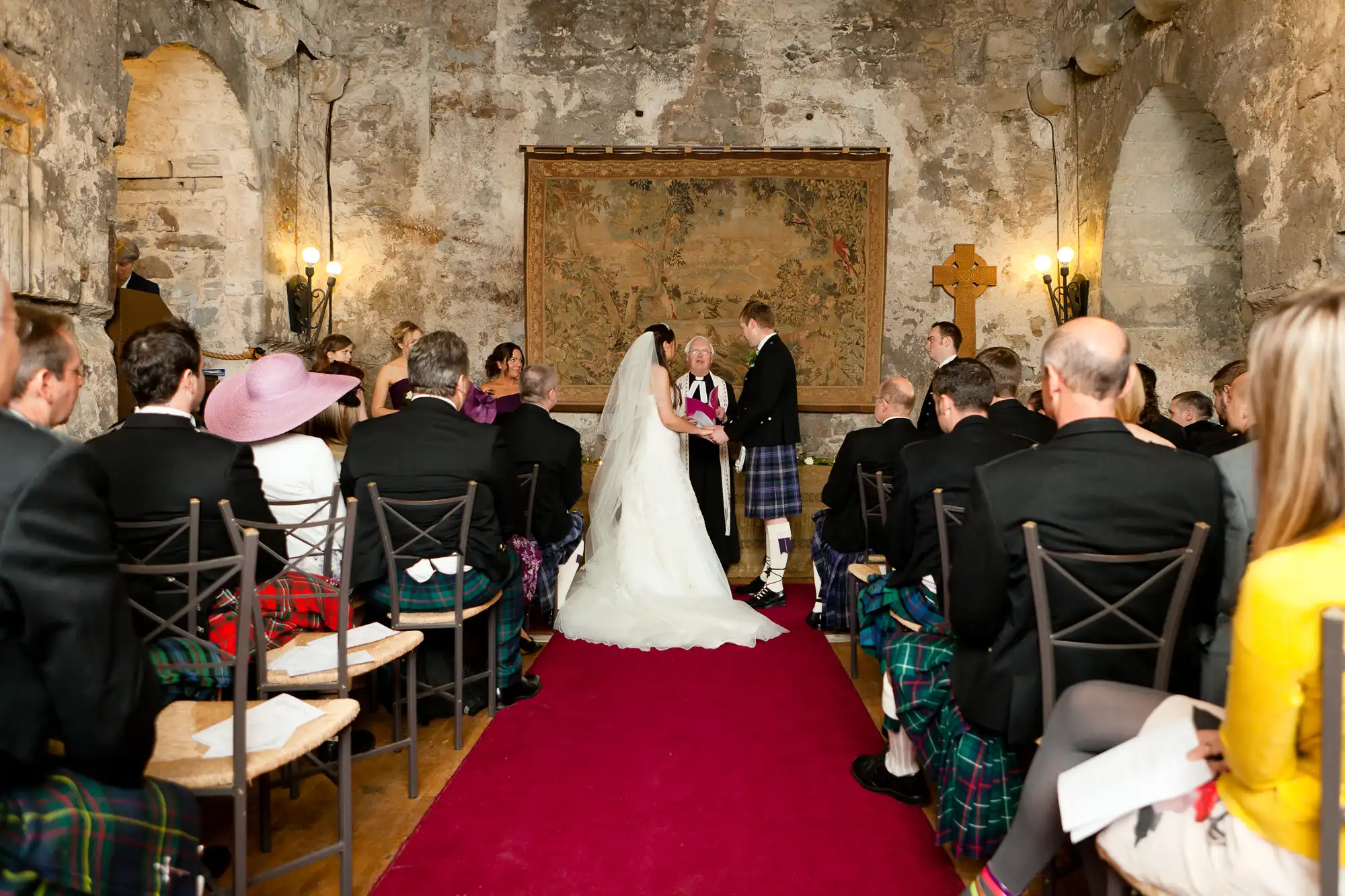 A bride and groom holding hands at the altar during a wedding ceremony in a rustic chapel, with guests in formal attire, some wearing kilts.
