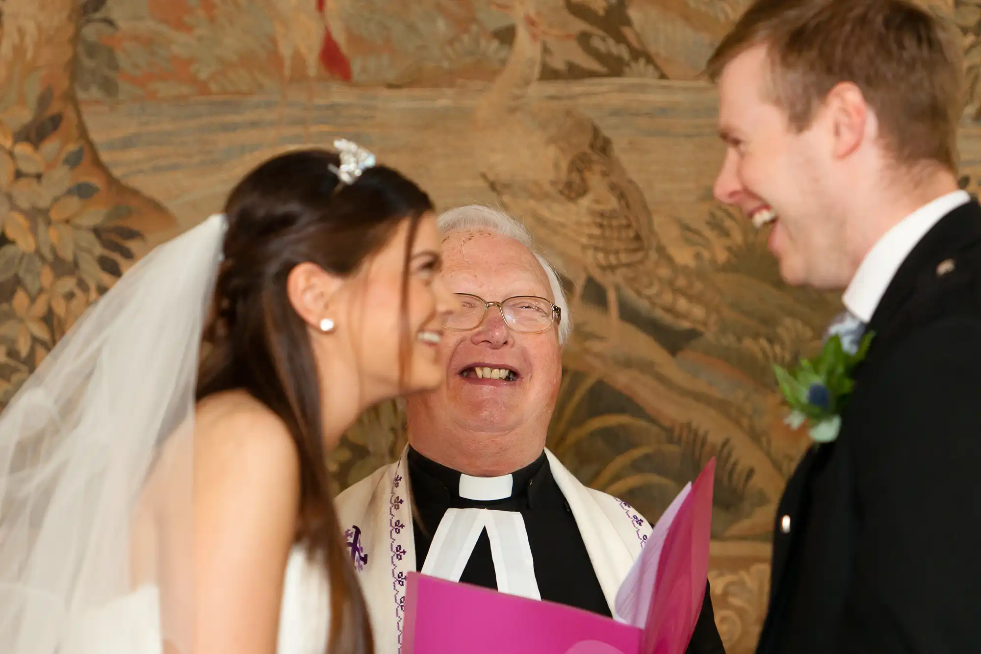 A bride and groom smiling at each other, with a joyful priest holding a pink booklet, during a wedding ceremony.