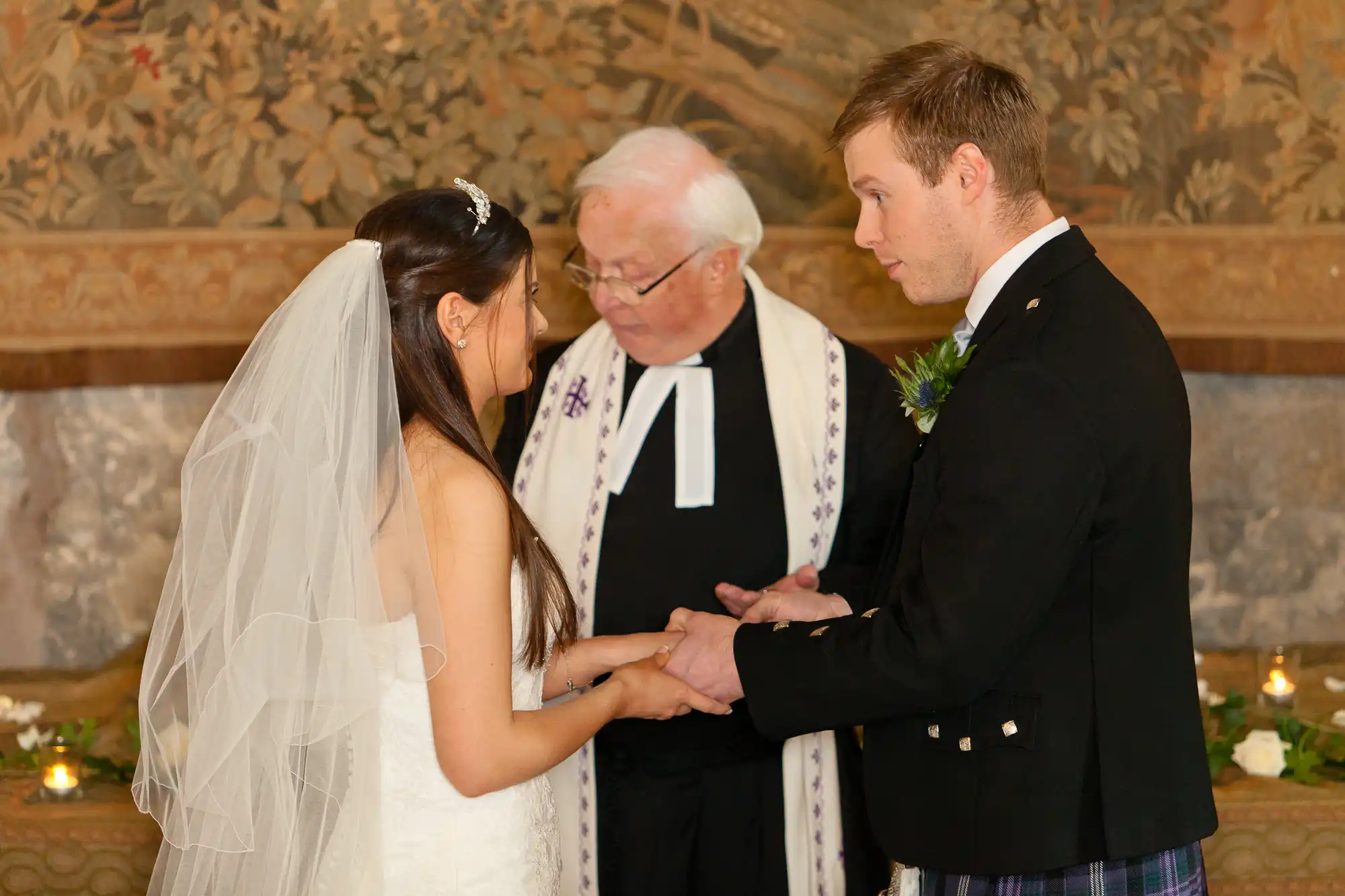 A bride and groom exchanging rings during a wedding ceremony, officiated by a priest, in a room with a mural in the background.