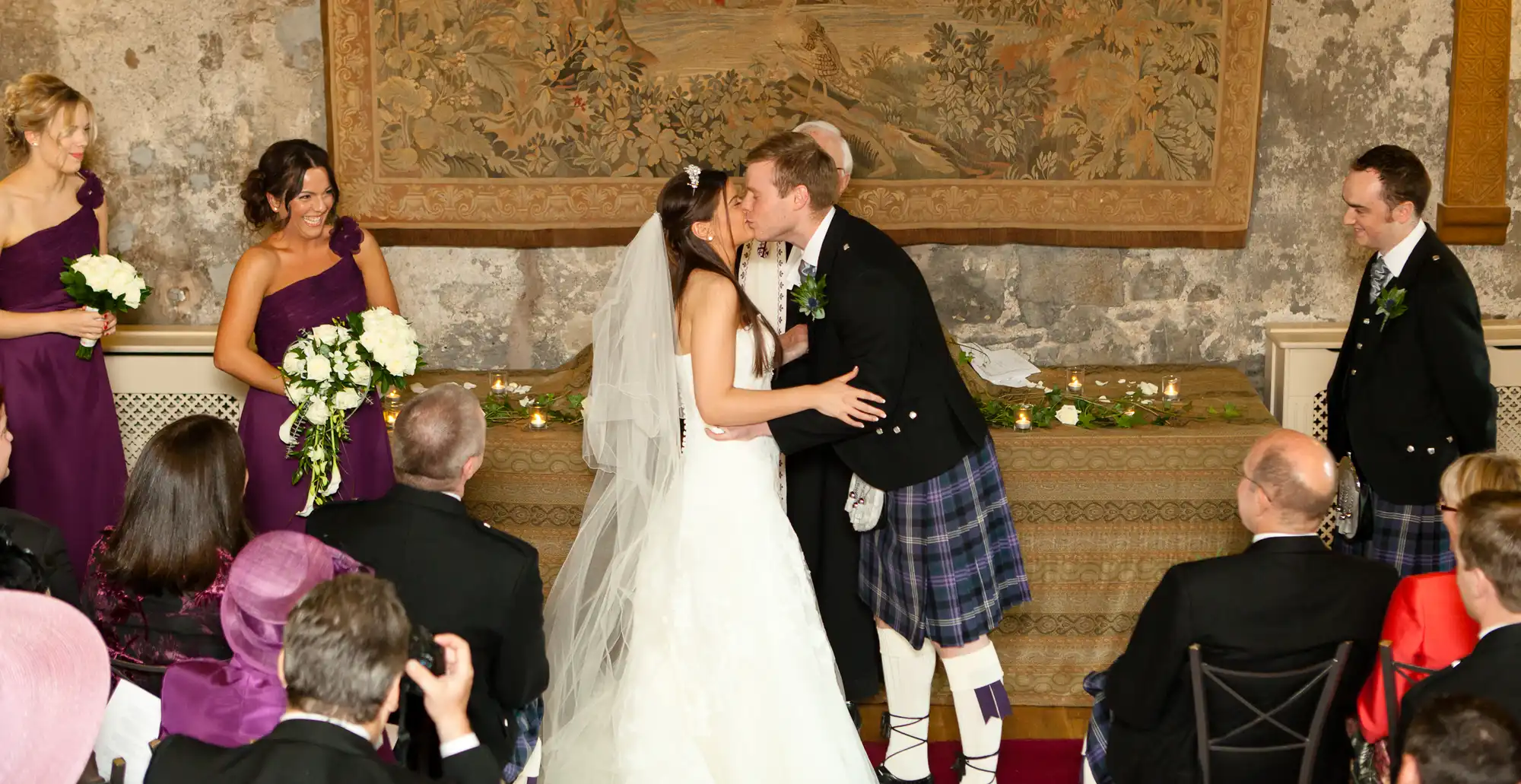 A bride and groom kissing at their wedding ceremony, with the groom wearing a kilt. bridesmaids in purple dresses and guests are watching.