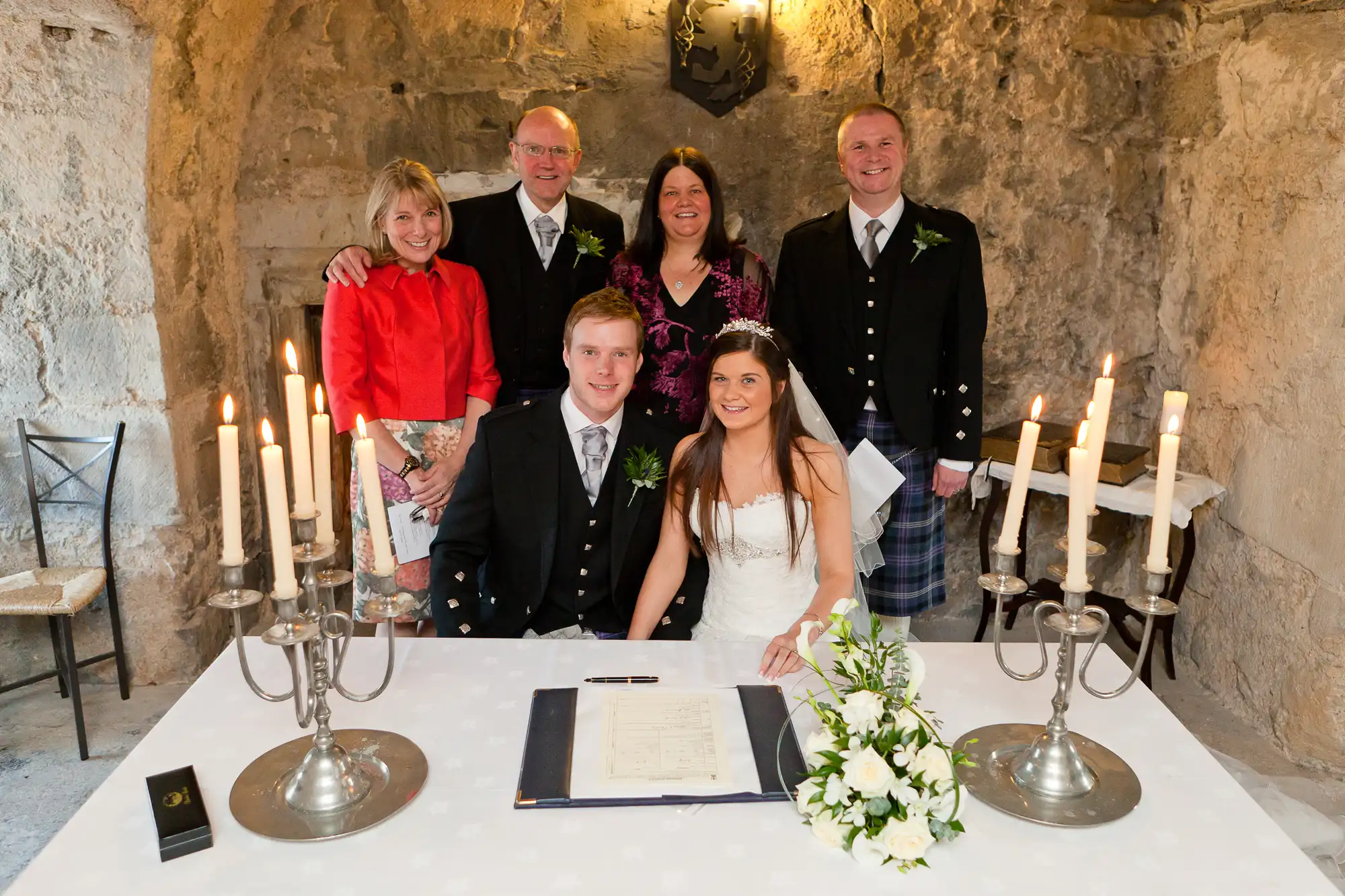 A newlywed couple signing a marriage register at a table adorned with candles and flowers, accompanied by four smiling guests in formal attire.