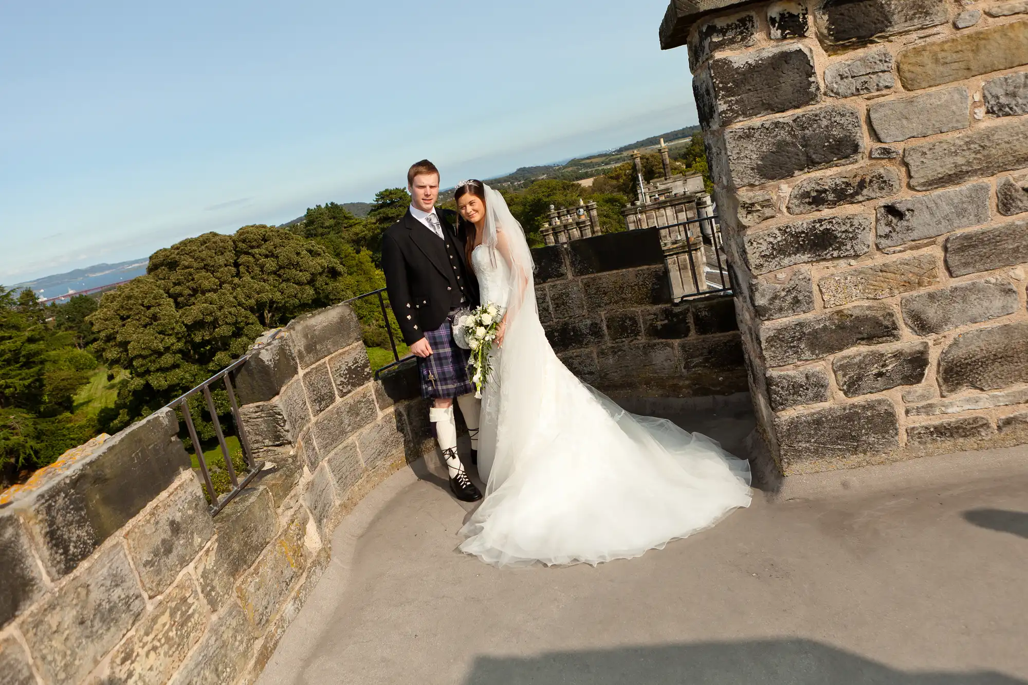 A newlywed couple in wedding attire, the groom in a kilt, stands on a stone castle walkway, with scenic trees and water in the background.