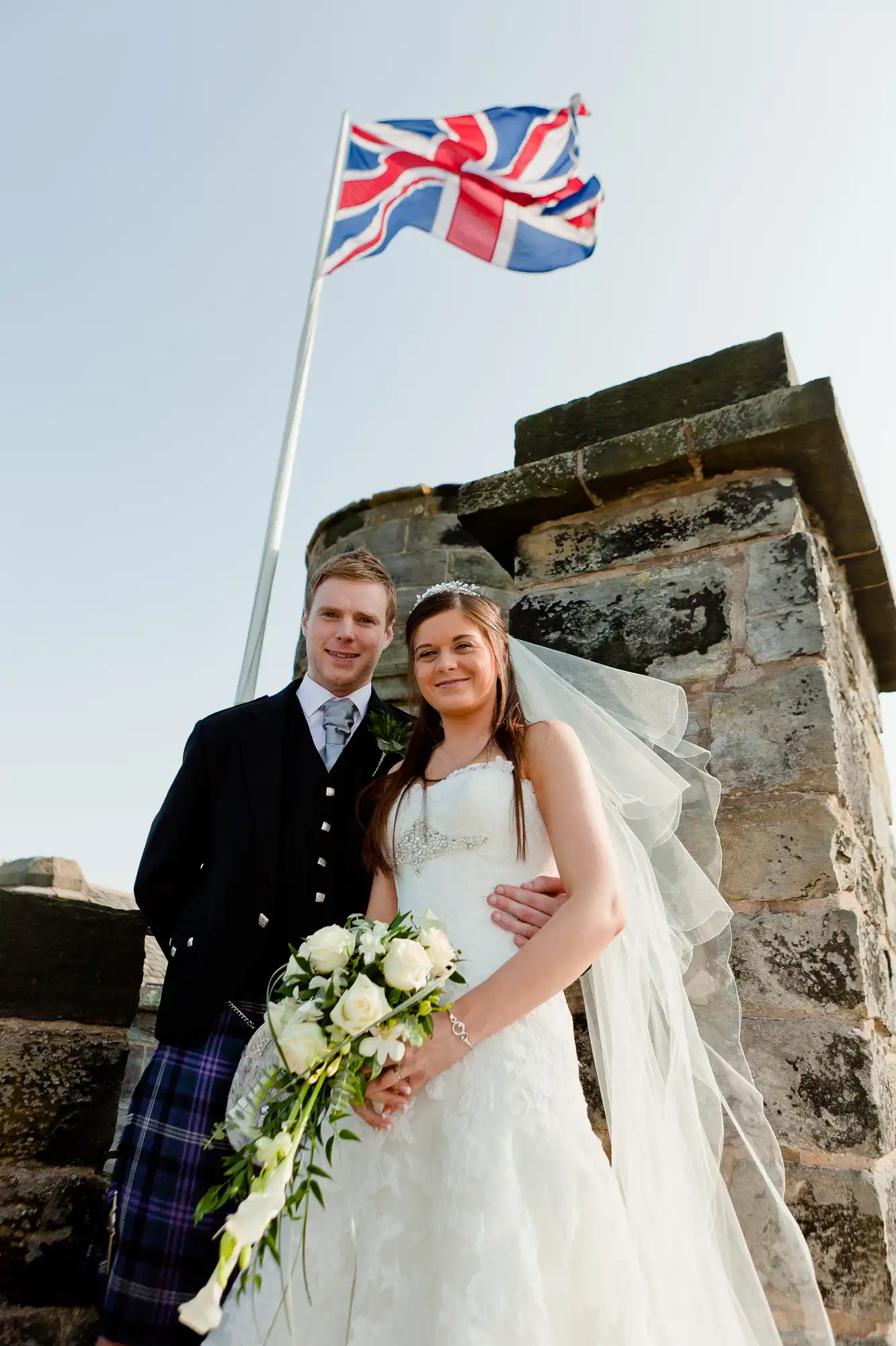 A newlywed couple posing under a british flag at a historic castle, with the groom in tartan kilt and bride holding a bouquet.