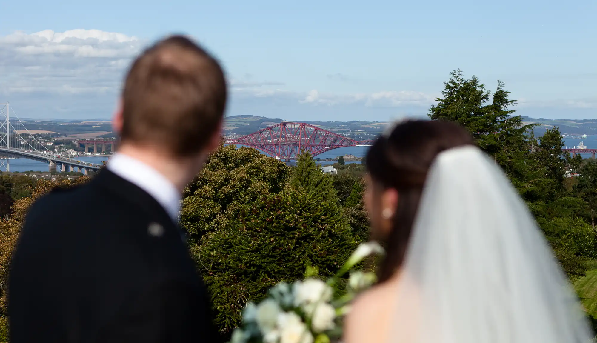 A bride and groom, viewed from behind, overlook a scenic view featuring the forth bridge and lush greenery.