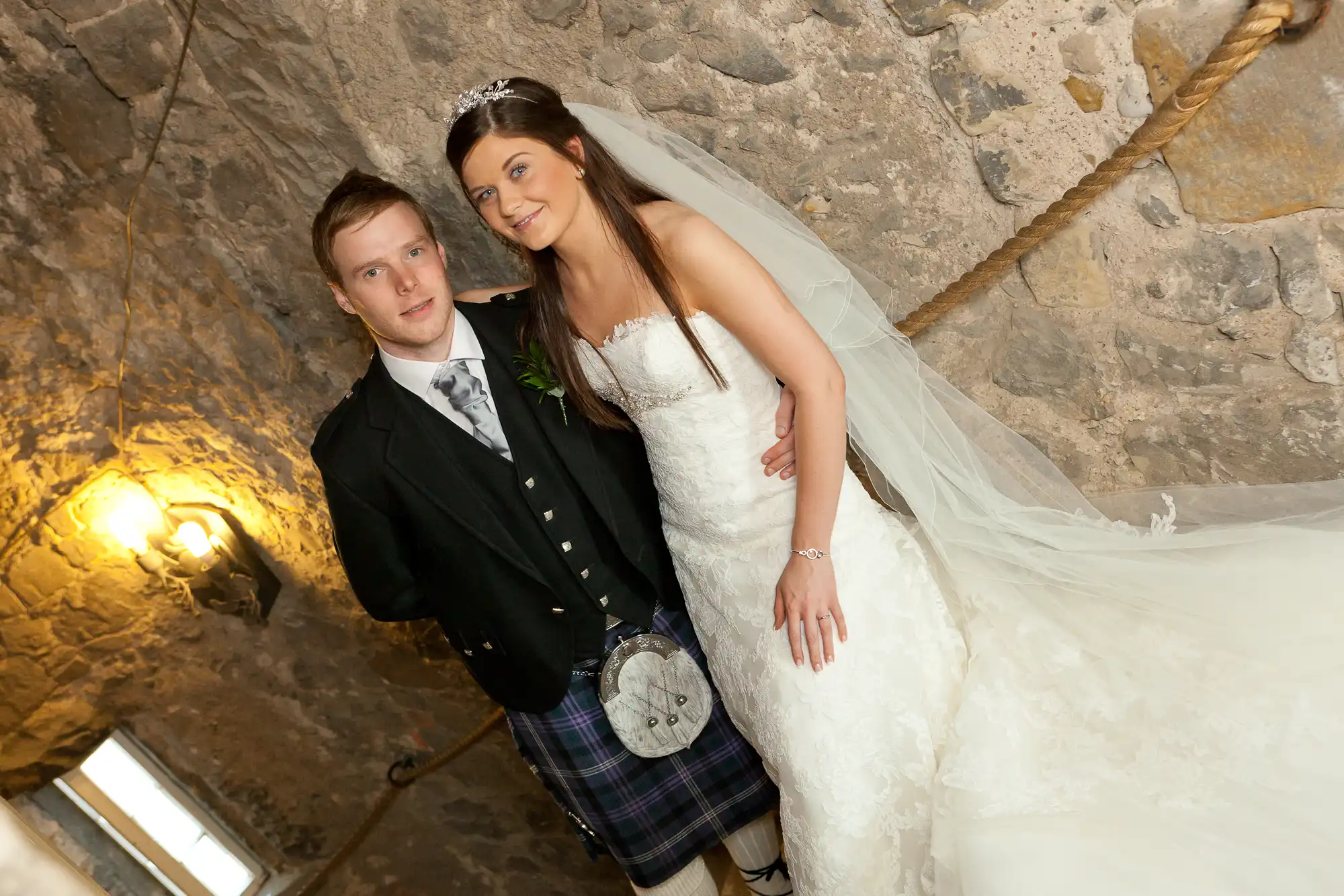 A newlywed couple posing in a stone-walled room, the groom in a kilt and the bride in a white dress, both smiling at the camera.