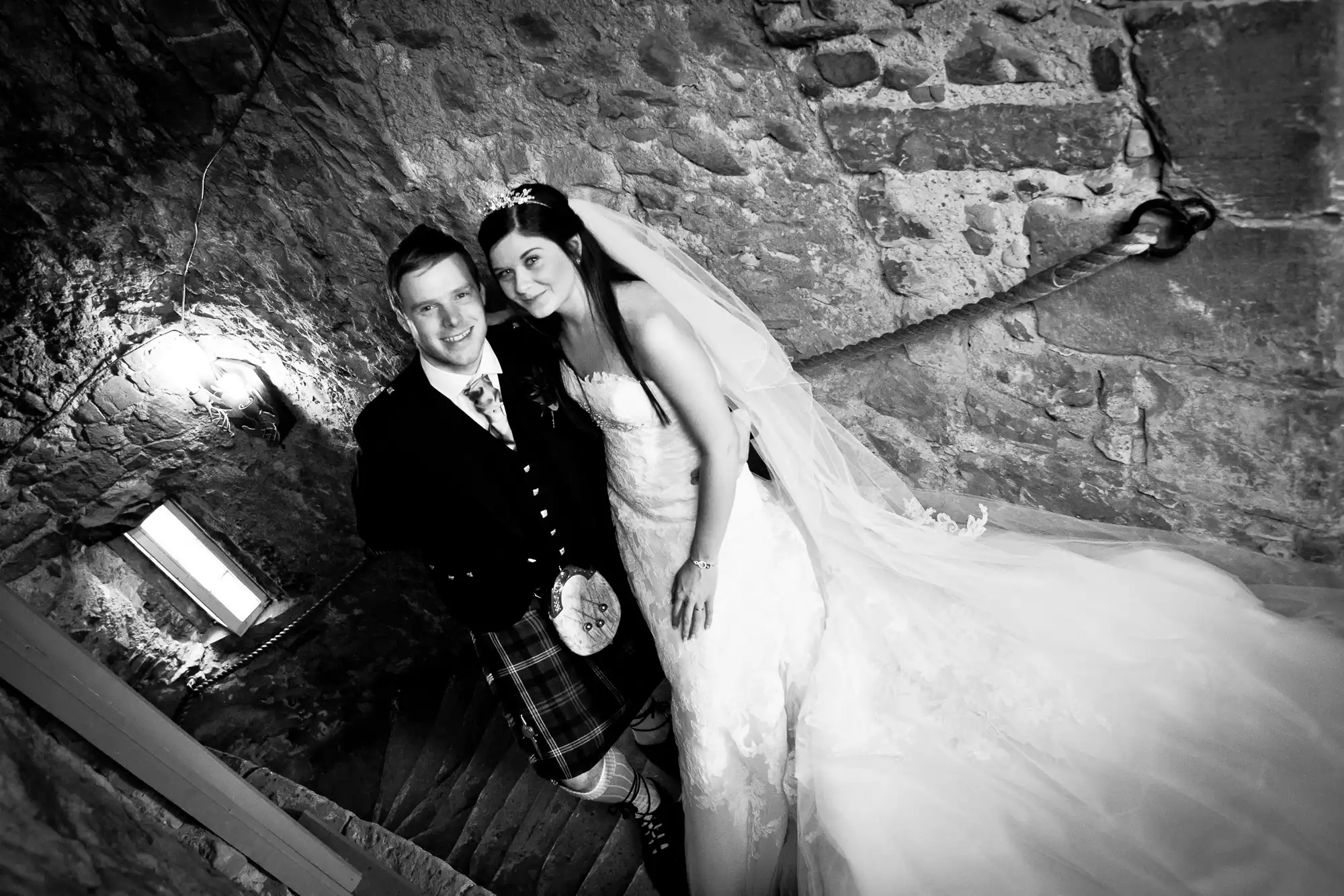 A bride and groom in wedding attire, the groom in a kilt, smiling inside a stone-walled stairwell, creating a romantic atmosphere.