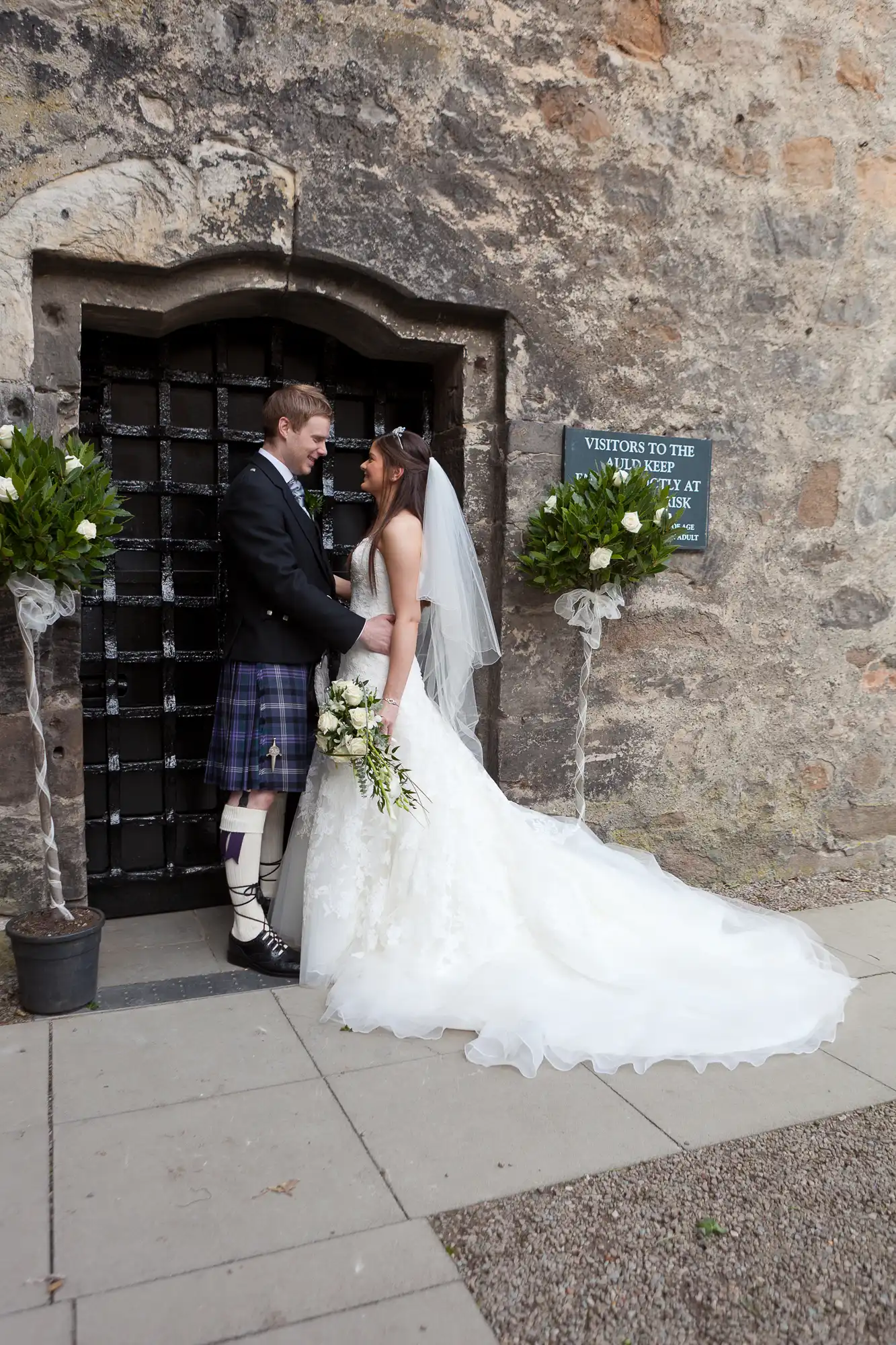 Bride in a white dress and groom in a kilt standing by a stone building with a gated doorway, sharing a moment.