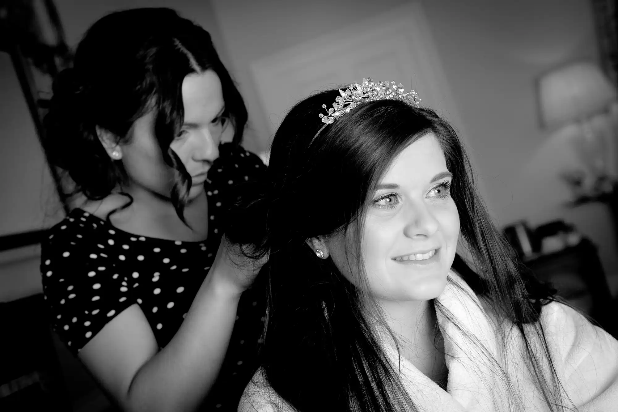 A woman in a white robe and tiara smiles while another woman styles her hair in a room with soft lighting.