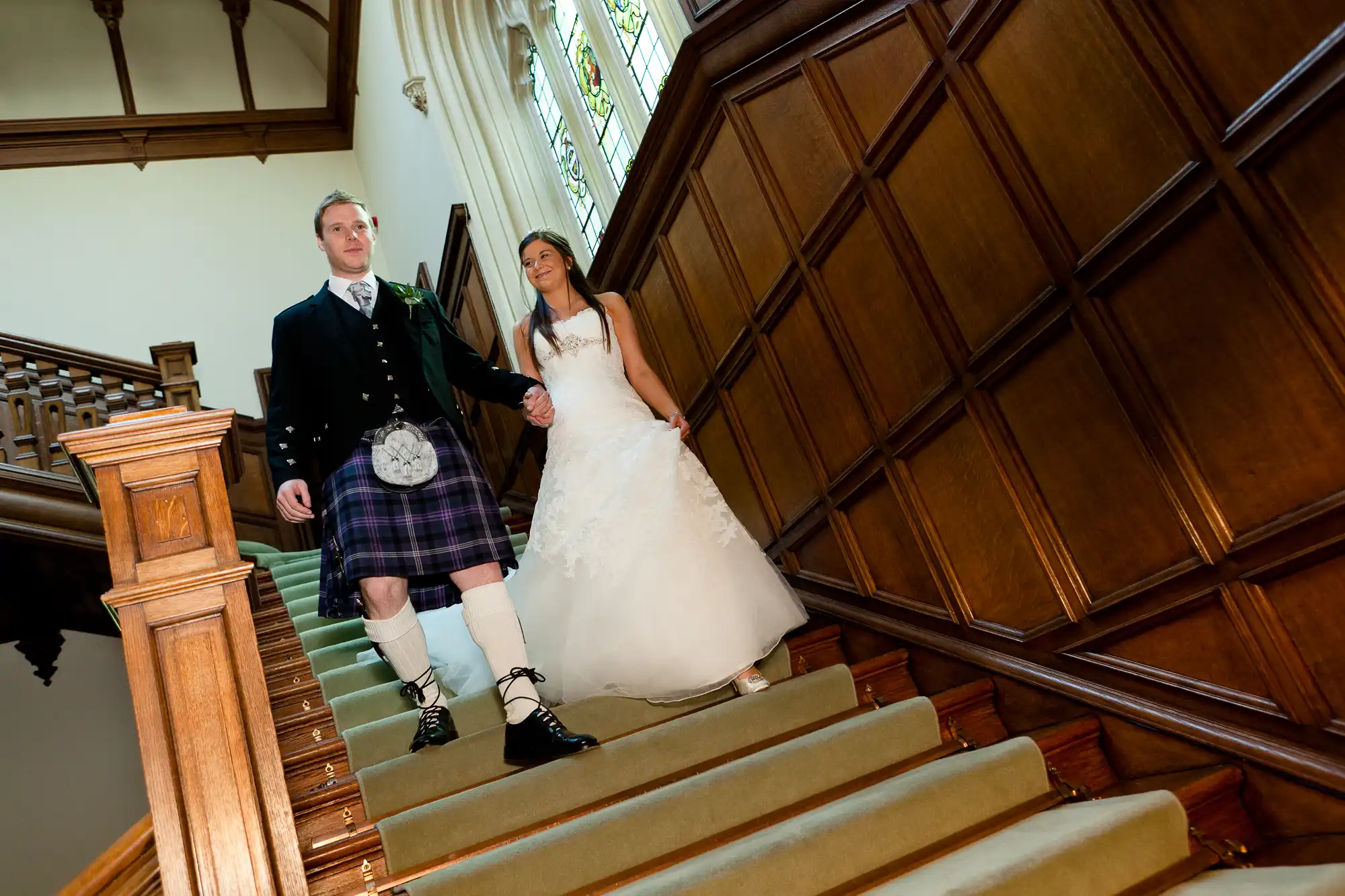 A bride in a white dress and a groom in a kilt holding hands while descending a staircase in a grand wood-paneled hall.