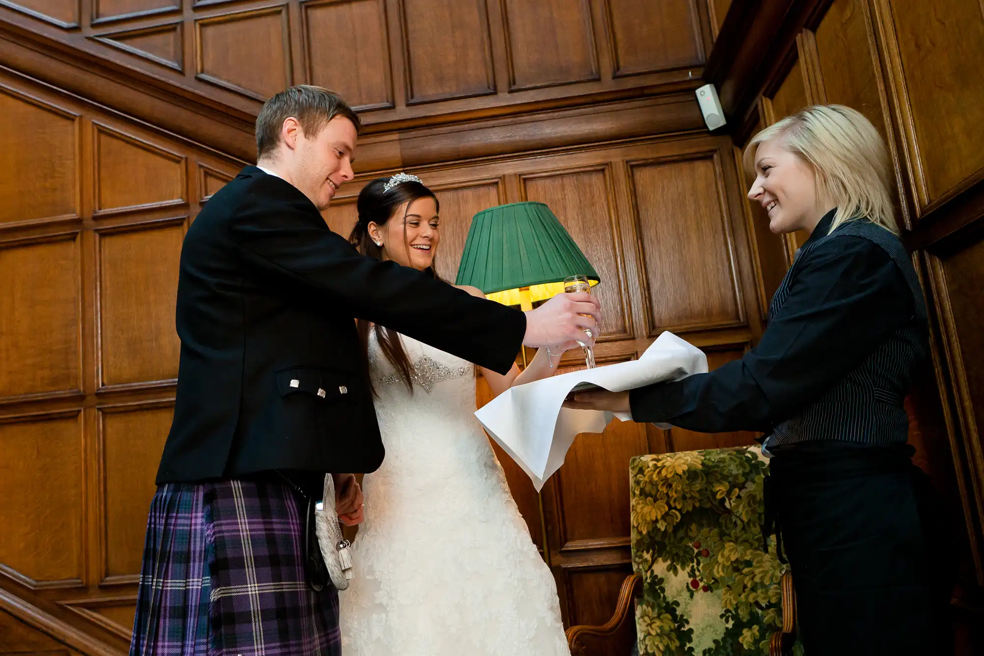 A bride and groom in a kilt and white dress laugh while signing their marriage certificate, handed by a smiling female officiant in a dark suit.