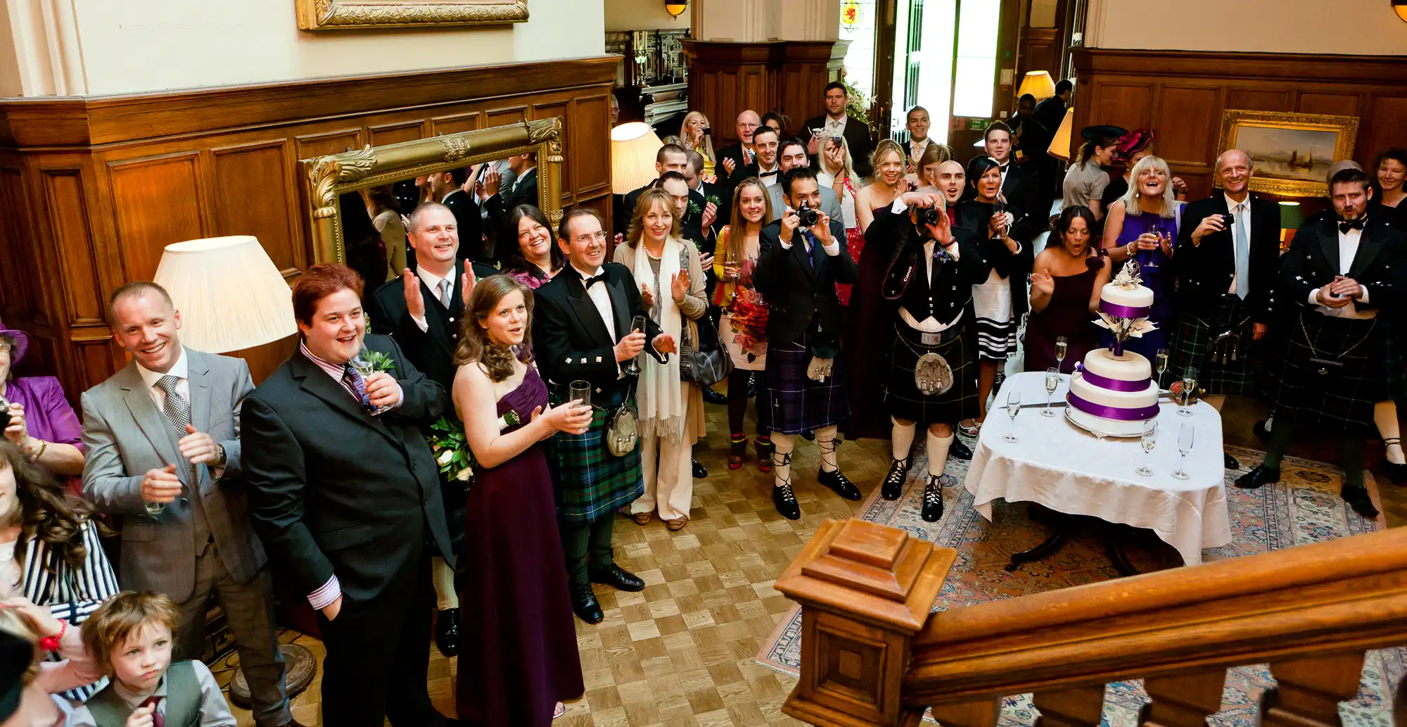 A diverse group of people in formal attire, including kilts, cheer with drinks at a wedding reception inside an elegant room.