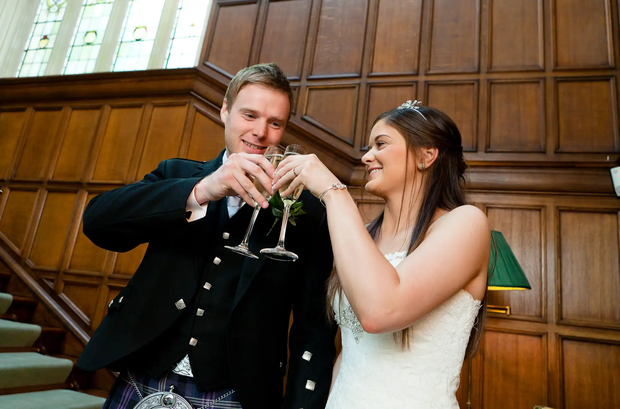 A bride and groom in wedding attire, clinking glasses in a toast, smiling in a room with wood paneling and stained glass windows.