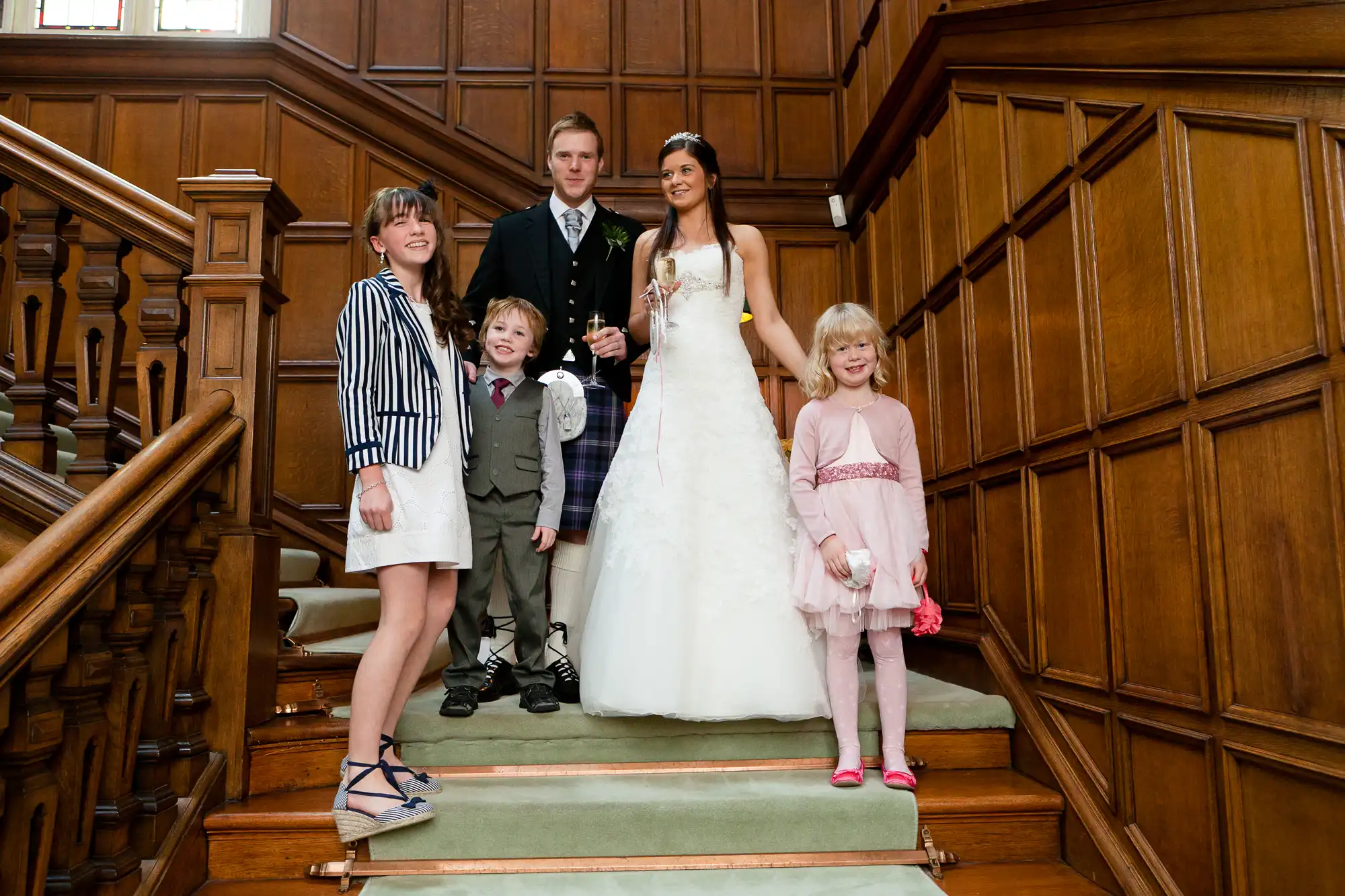 A newlywed couple standing on a staircase with three children, inside a vintage wooden paneled room, smiling for a photo.