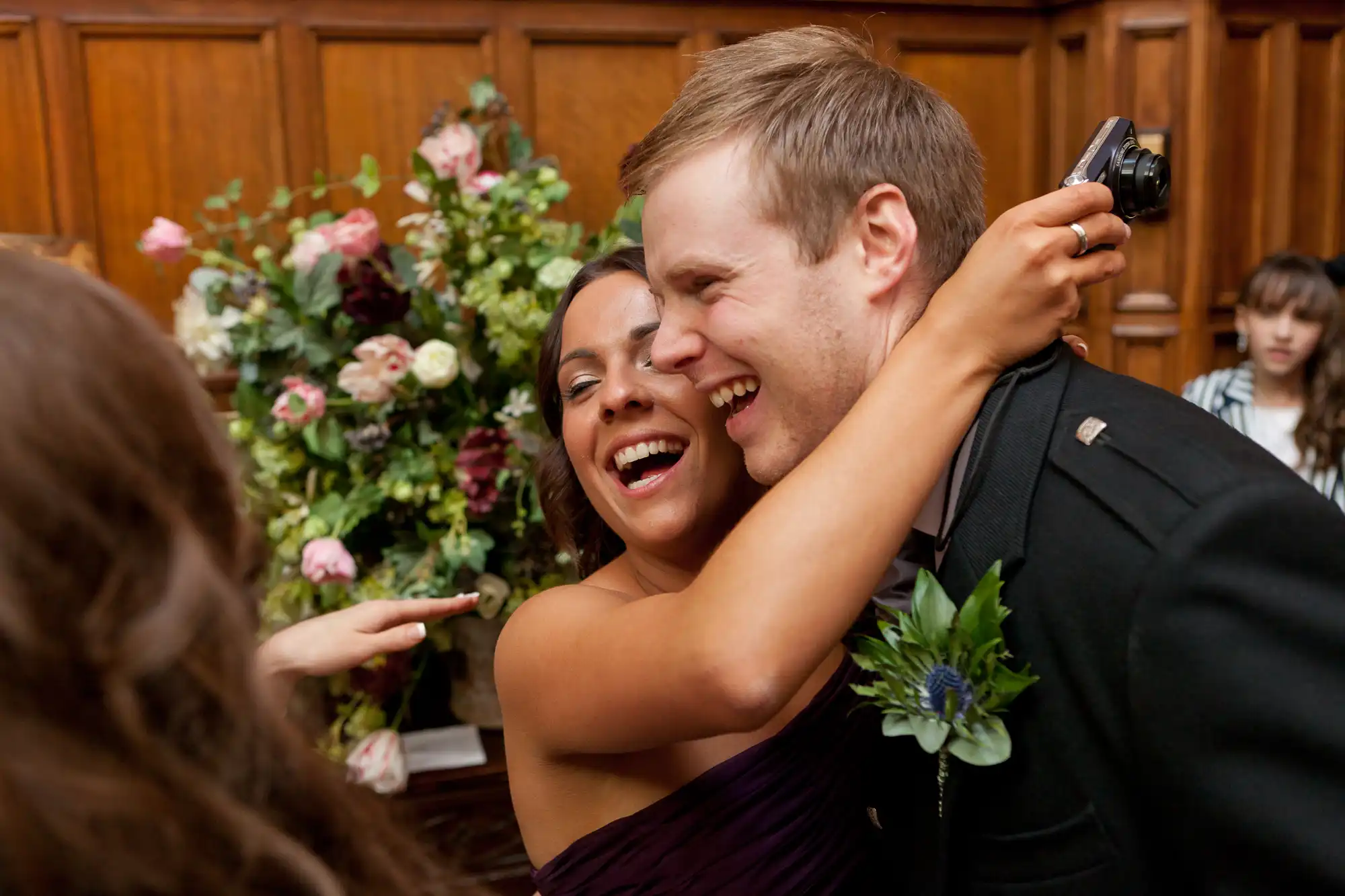 A joyful couple hugging and laughing at a wedding reception, surrounded by guests and floral decorations.