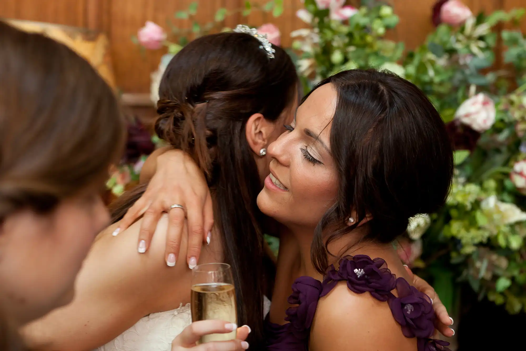 A bride and a bridesmaid embracing at a wedding reception, surrounded by floral decorations. the bridesmaid holds a champagne glass.