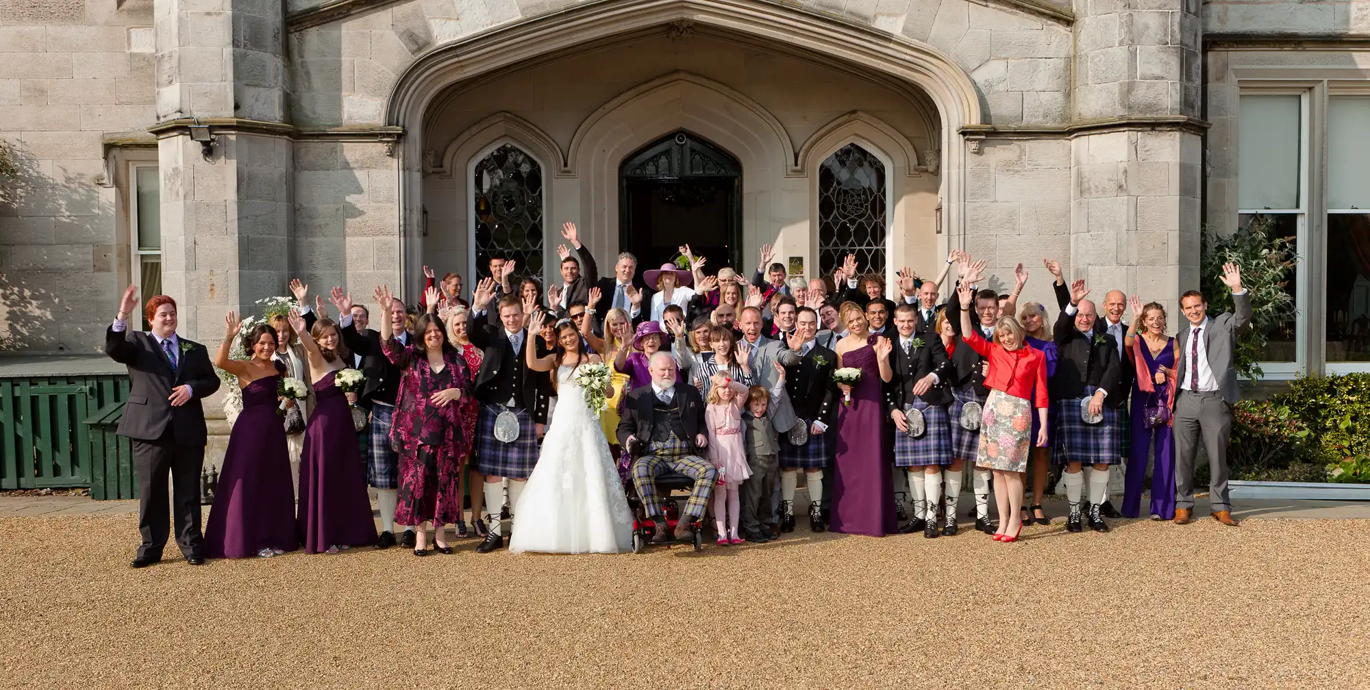 A large group of wedding guests celebrating outside a grand stone building, with the bride in a white dress and attendees in formal attire including kilts.