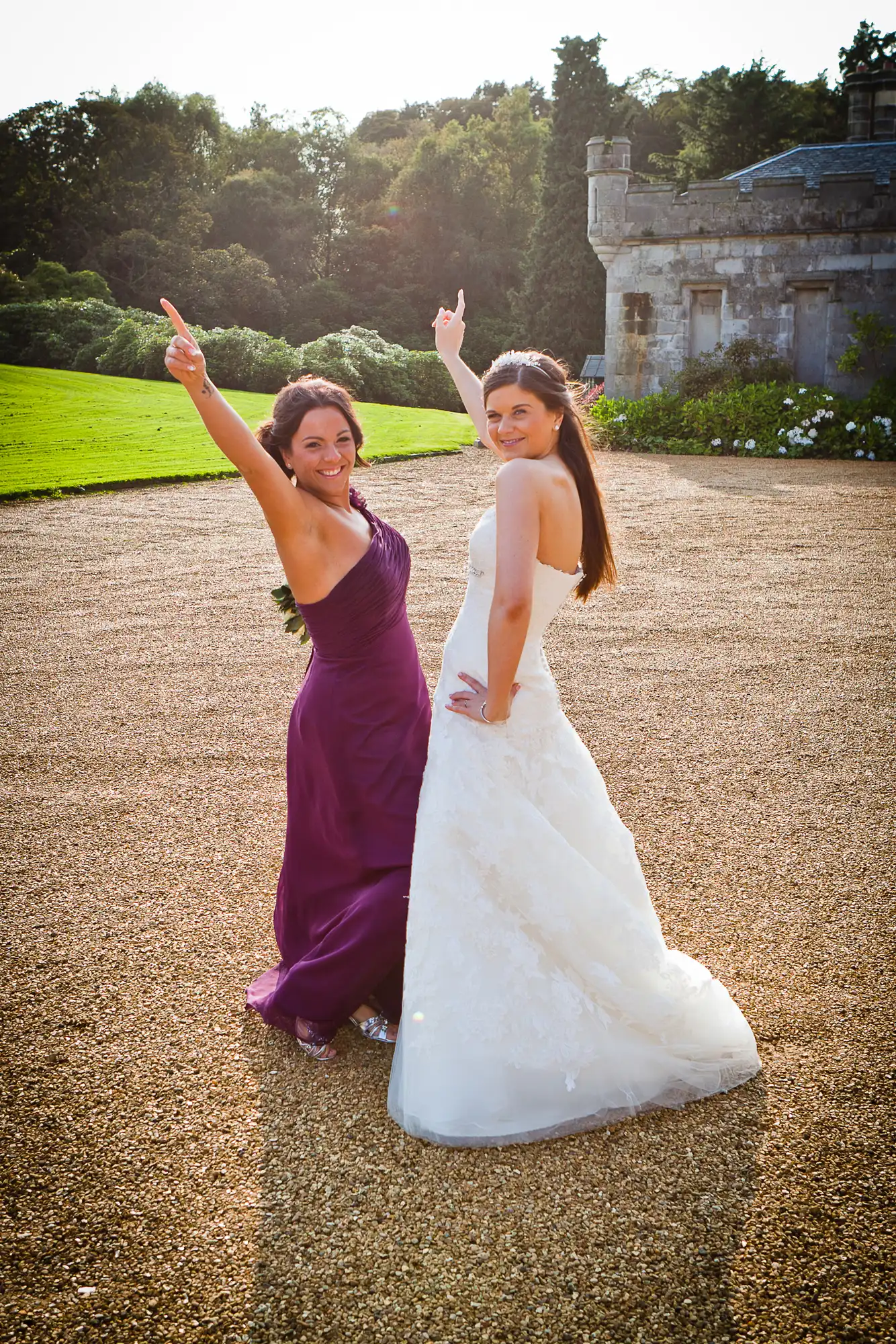A bride in a white gown and a bridesmaid in a purple dress joyously pose with their hands up in front of a stately home and green lawn.