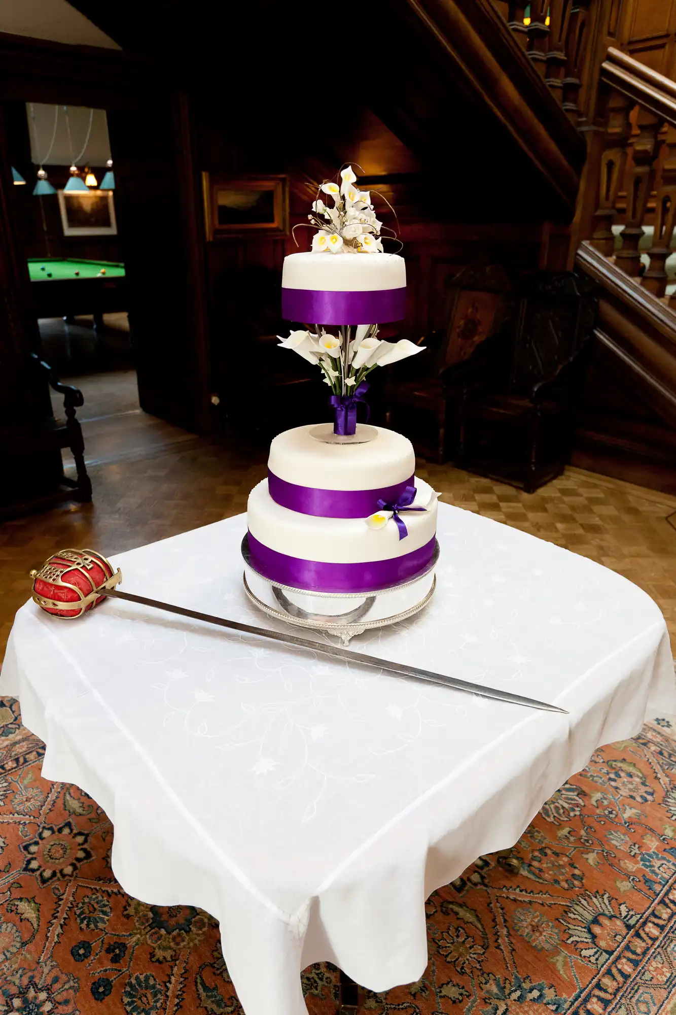 A three-tiered wedding cake with purple ribbons, decorated with white and purple flowers, displayed on a table in an elegant room with a wood staircase.