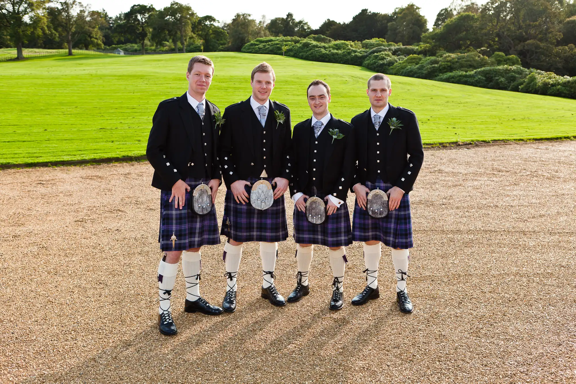 Four men in traditional scottish attire, including kilts and sporran, standing together on a grassy field with trees in the background.