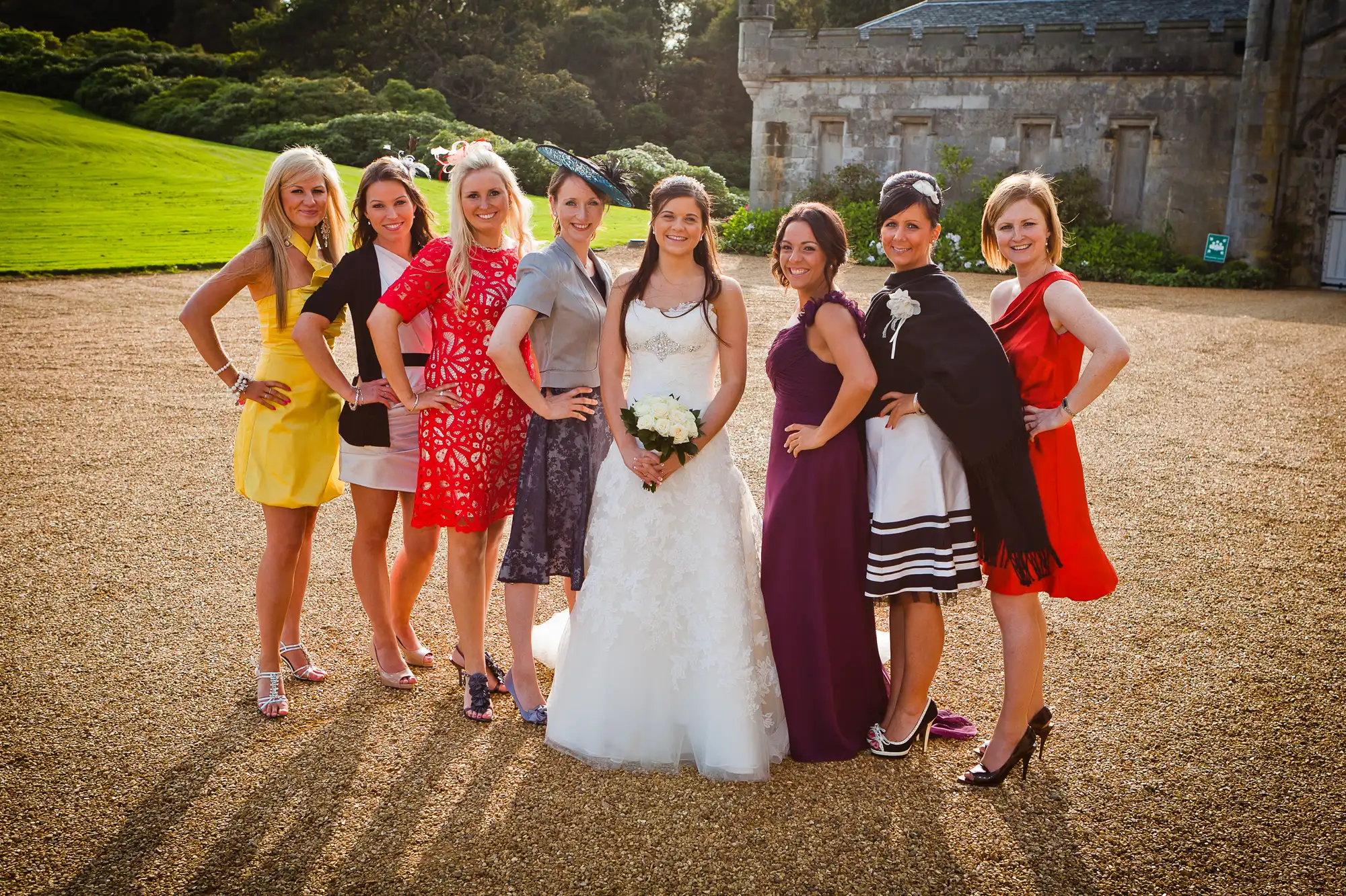 A bride in a white dress with six women in various colorful dresses posing together in front of a stately home on a sunny day.