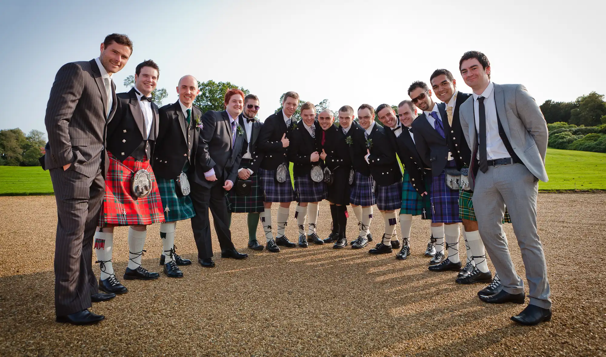 Group of men in formal attire and kilts posing outdoors on a sunny day, with trees and a clear sky in the background.