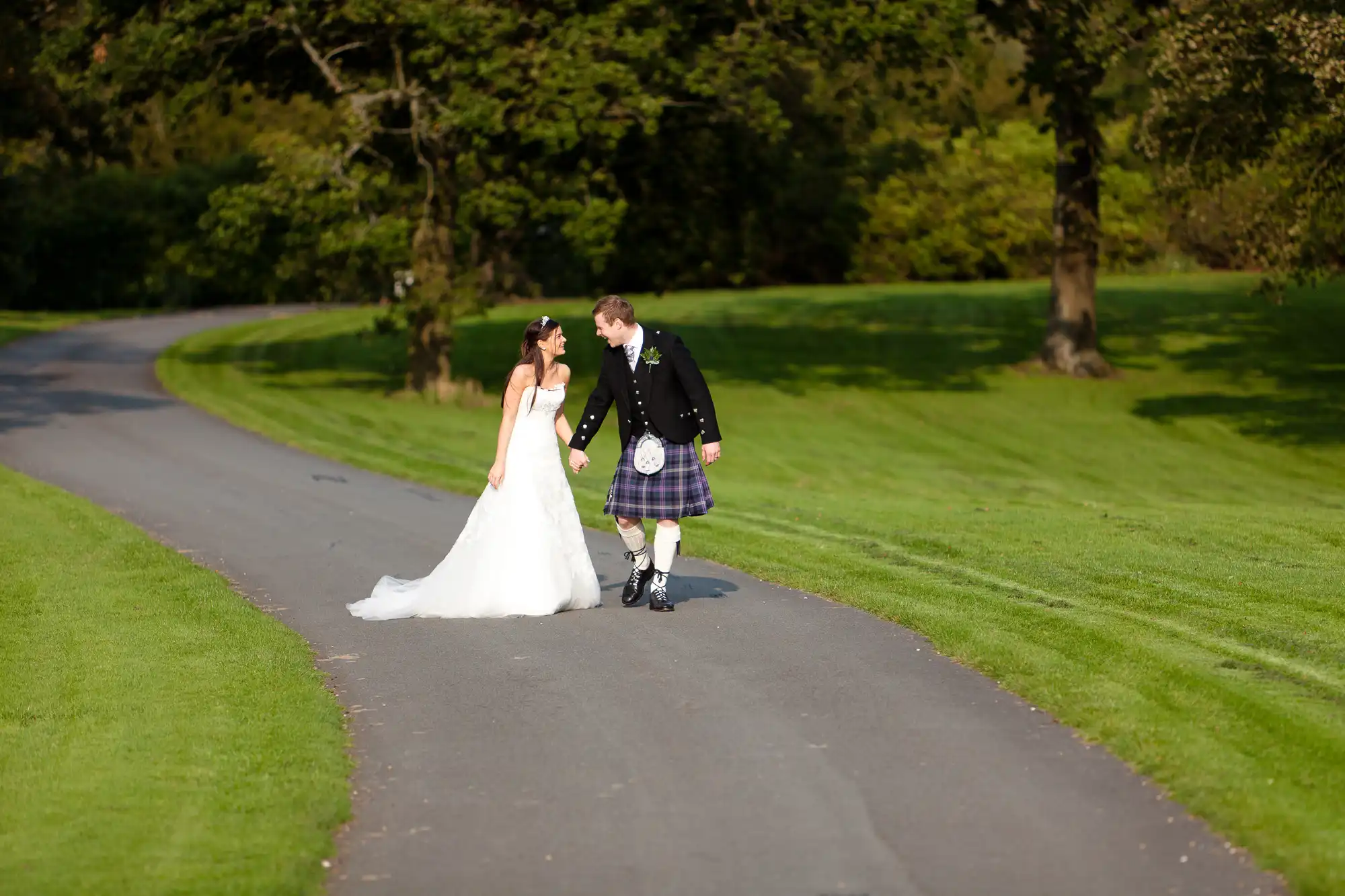 A bride in a white dress and a groom in a kilt walk hand in hand on a sunlit park pathway, sharing a kiss.