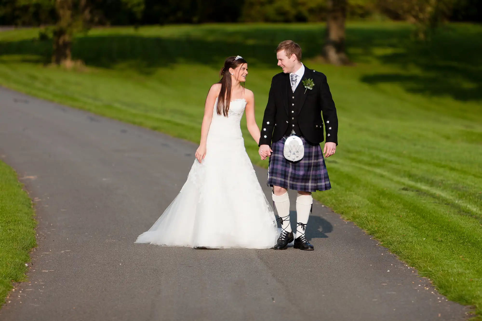 A bride in a white dress and a groom in a kilt walking and smiling at each other on a sunlit park pathway.
