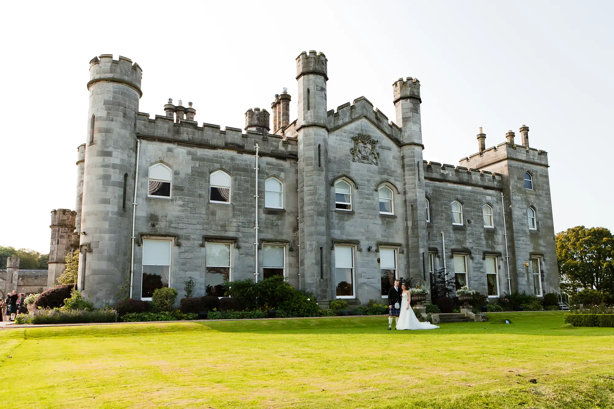 A bride and groom walk hand in hand on the lush lawn of an elegant, historic castle with tall towers.