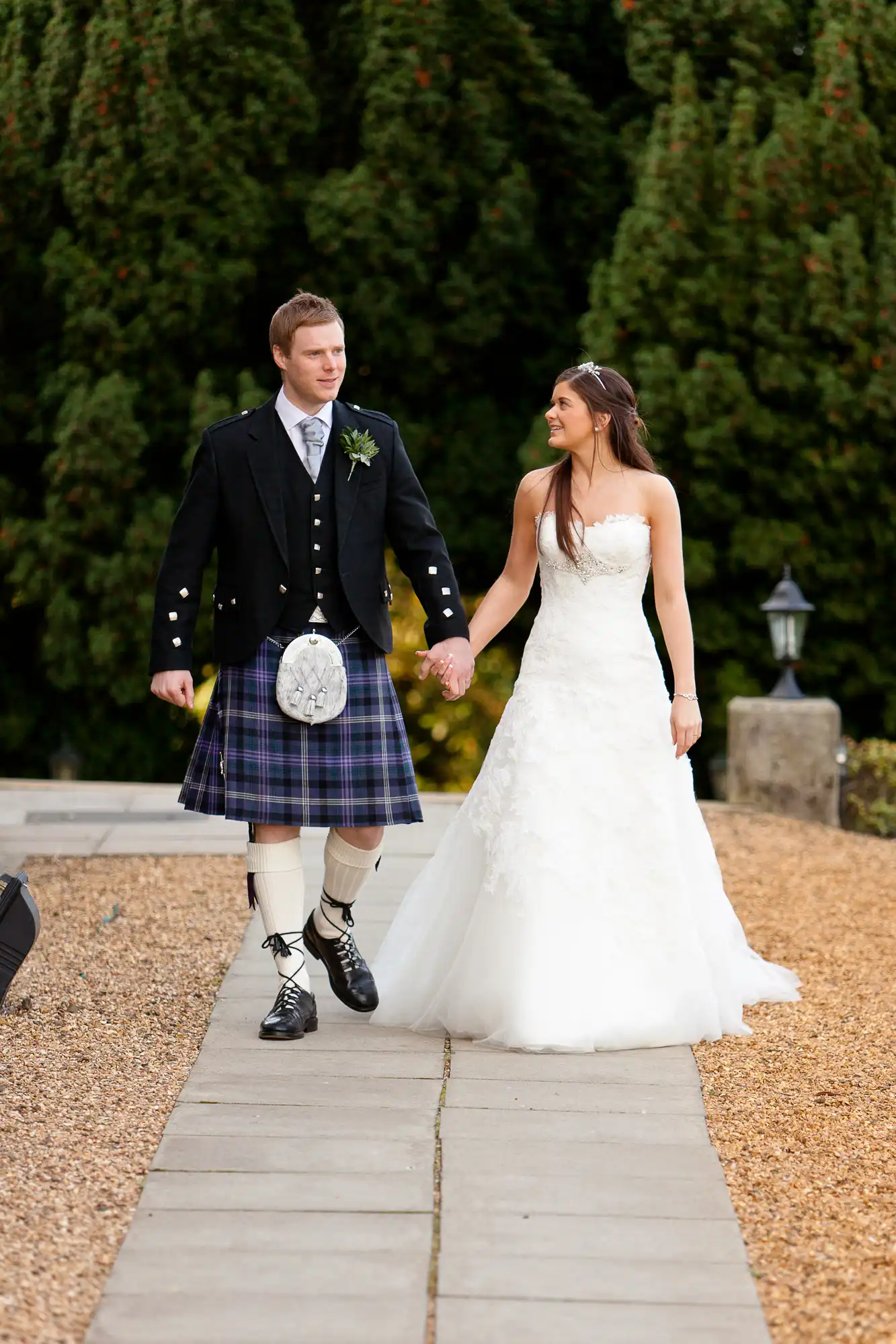 A bride in a white gown and a groom in a traditional scottish kilt and jacket walk hand in hand on a garden path.