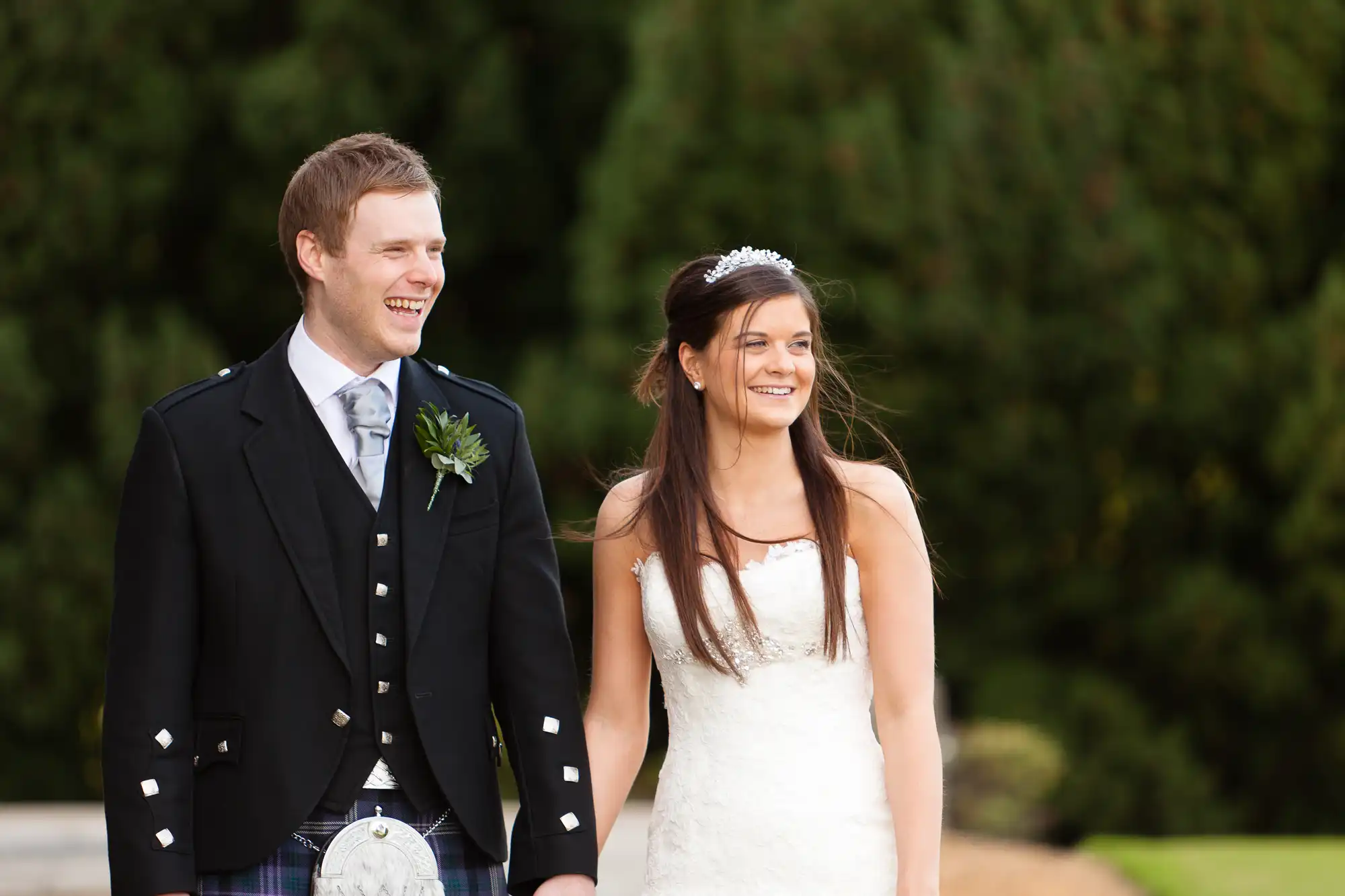 A bride and groom smiling and walking outdoors, the groom in a traditional kilt and the bride in a white dress.