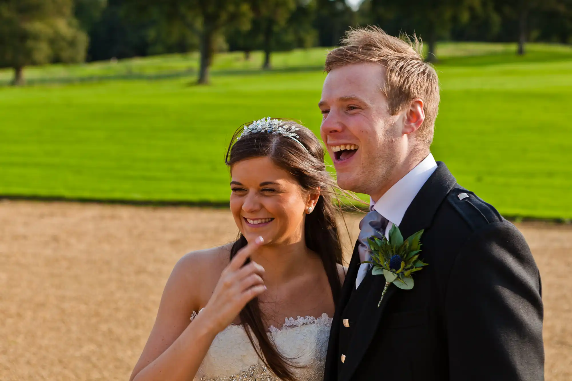 A bride and groom smiling brightly and walking outdoors on a sunny day, with the groom in a black suit and the bride in a white gown.