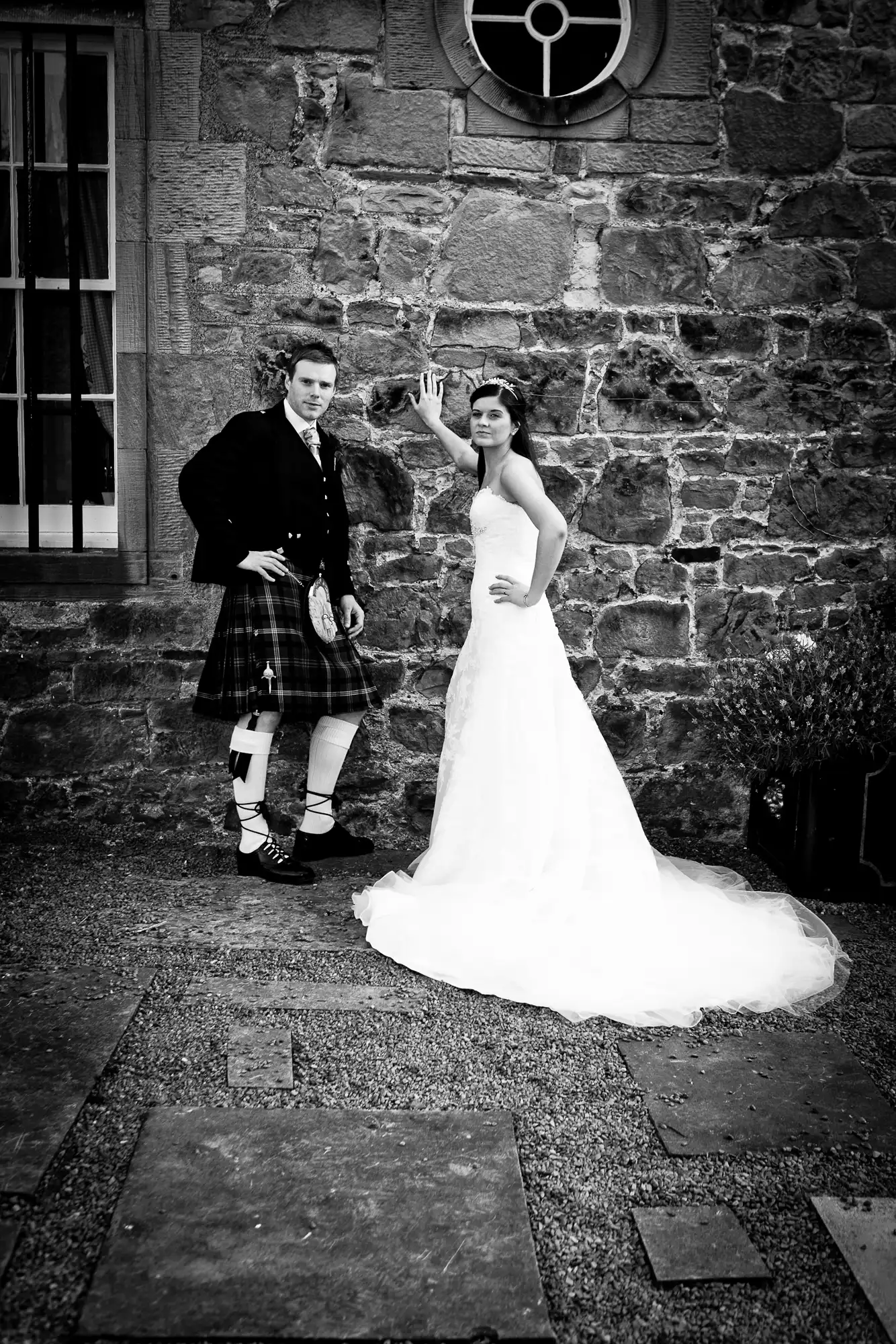 A bride and groom dressed in a white gown and a kilt wave to the camera in front of a stone building.