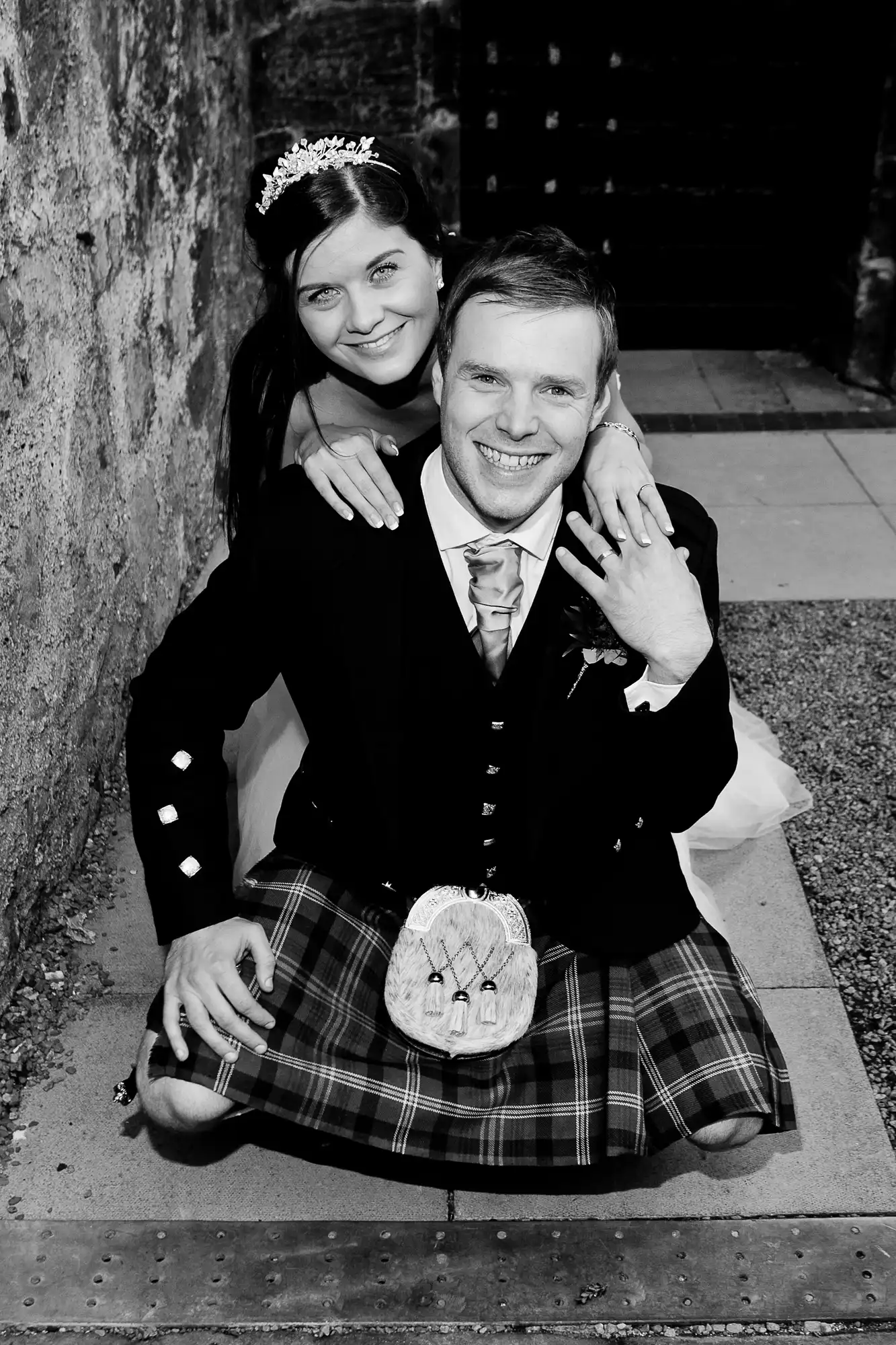A happy couple in wedding attire; the man wears a kilt and the woman, a tiara. they're seated closely, smiling, in a monochrome photo.