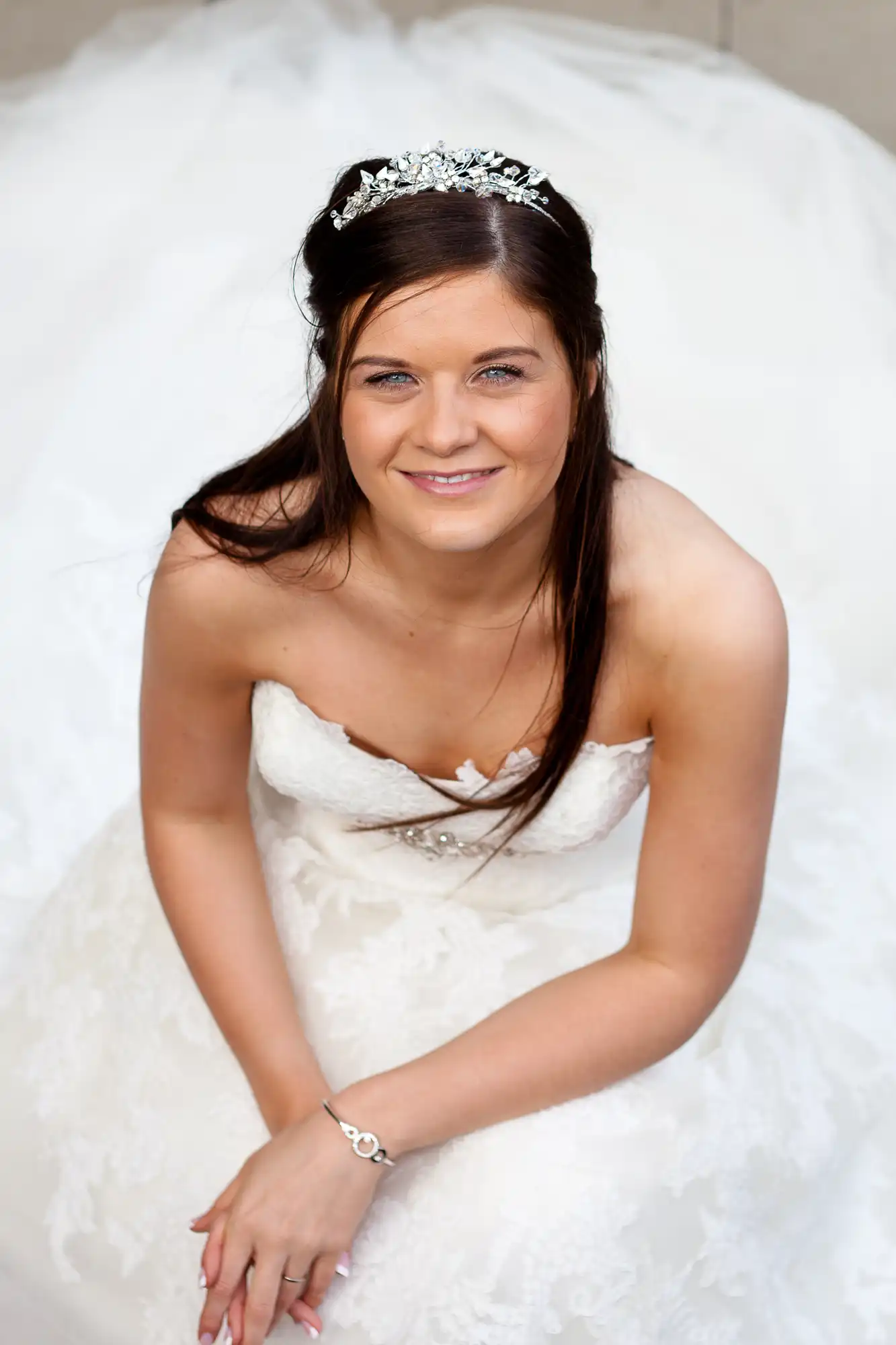 A smiling bride in a white gown and tiara sits elegantly, looking up at the camera.