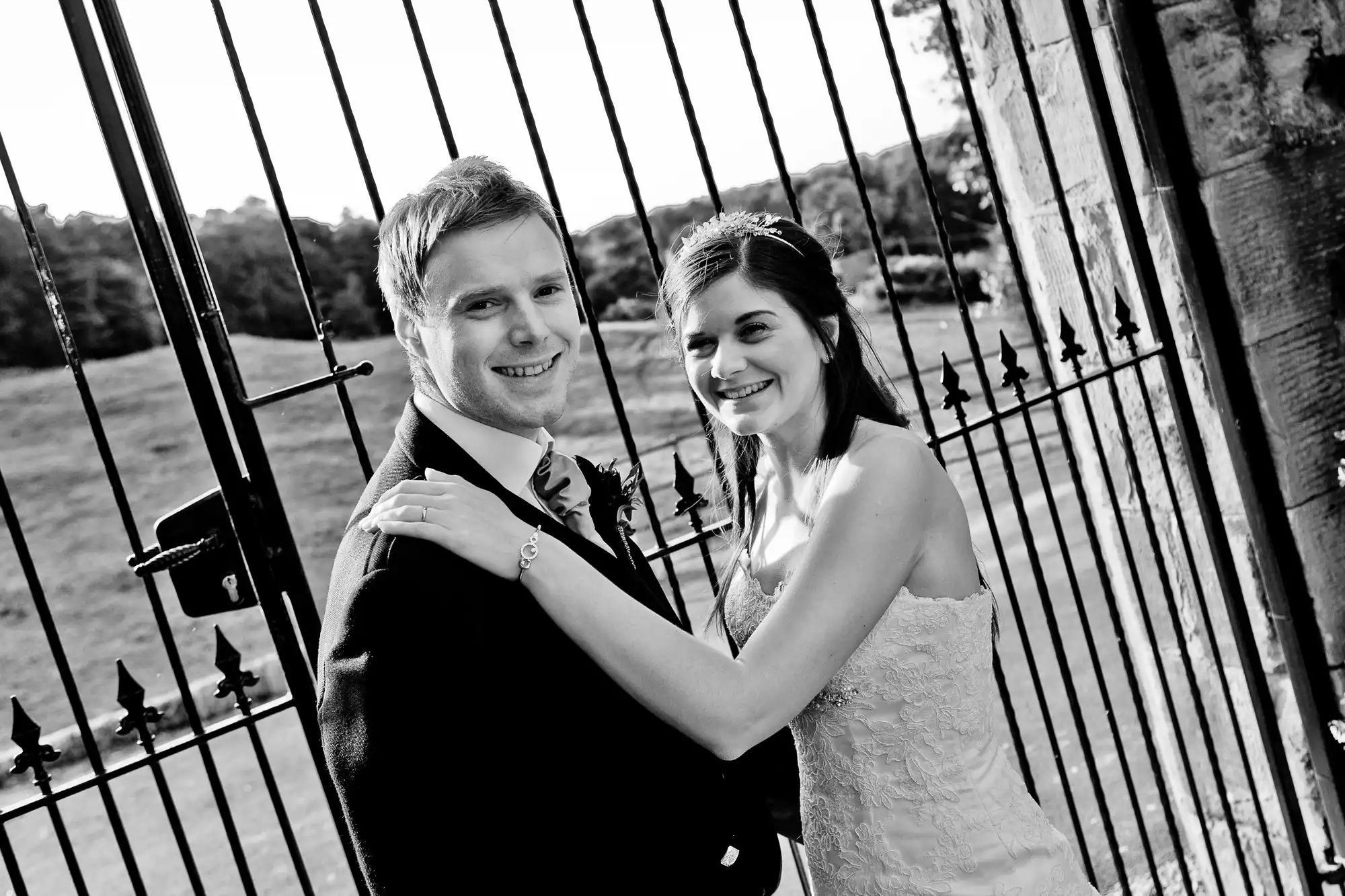A bride and groom smiling, embracing near a metal gate, in a black and white photograph.