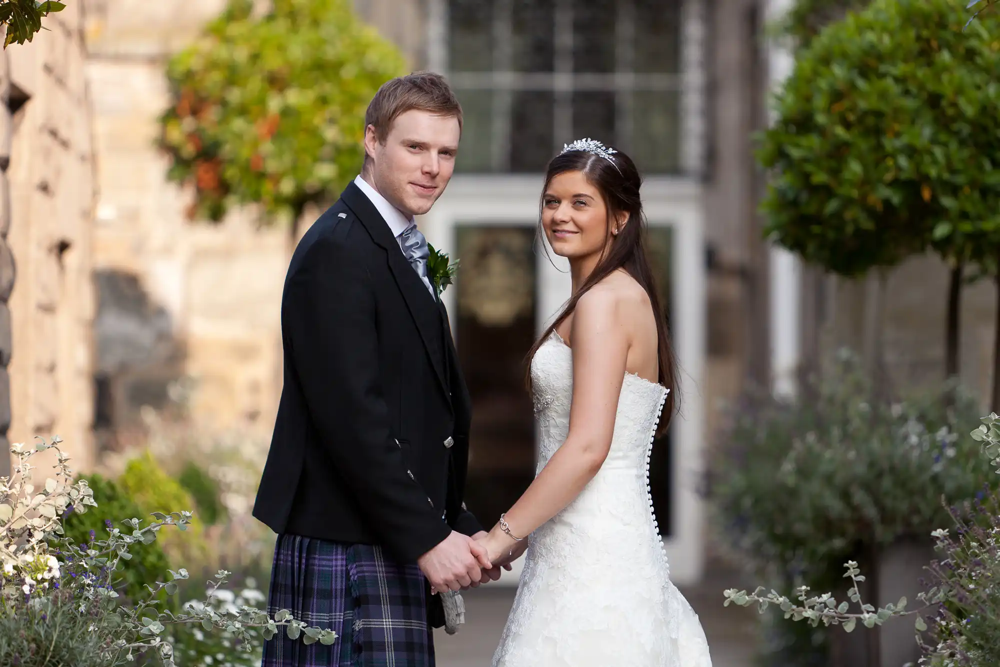 A bride and groom holding hands, with the groom in a kilt and the bride in a white dress, standing in a garden.