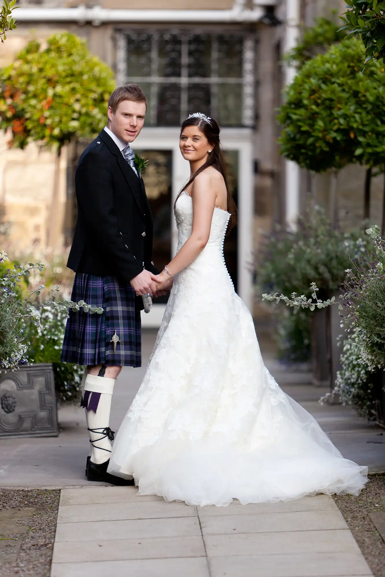 A bride in a white gown and a groom in a kilt holding hands in a garden pathway, with a historical building in the background.
