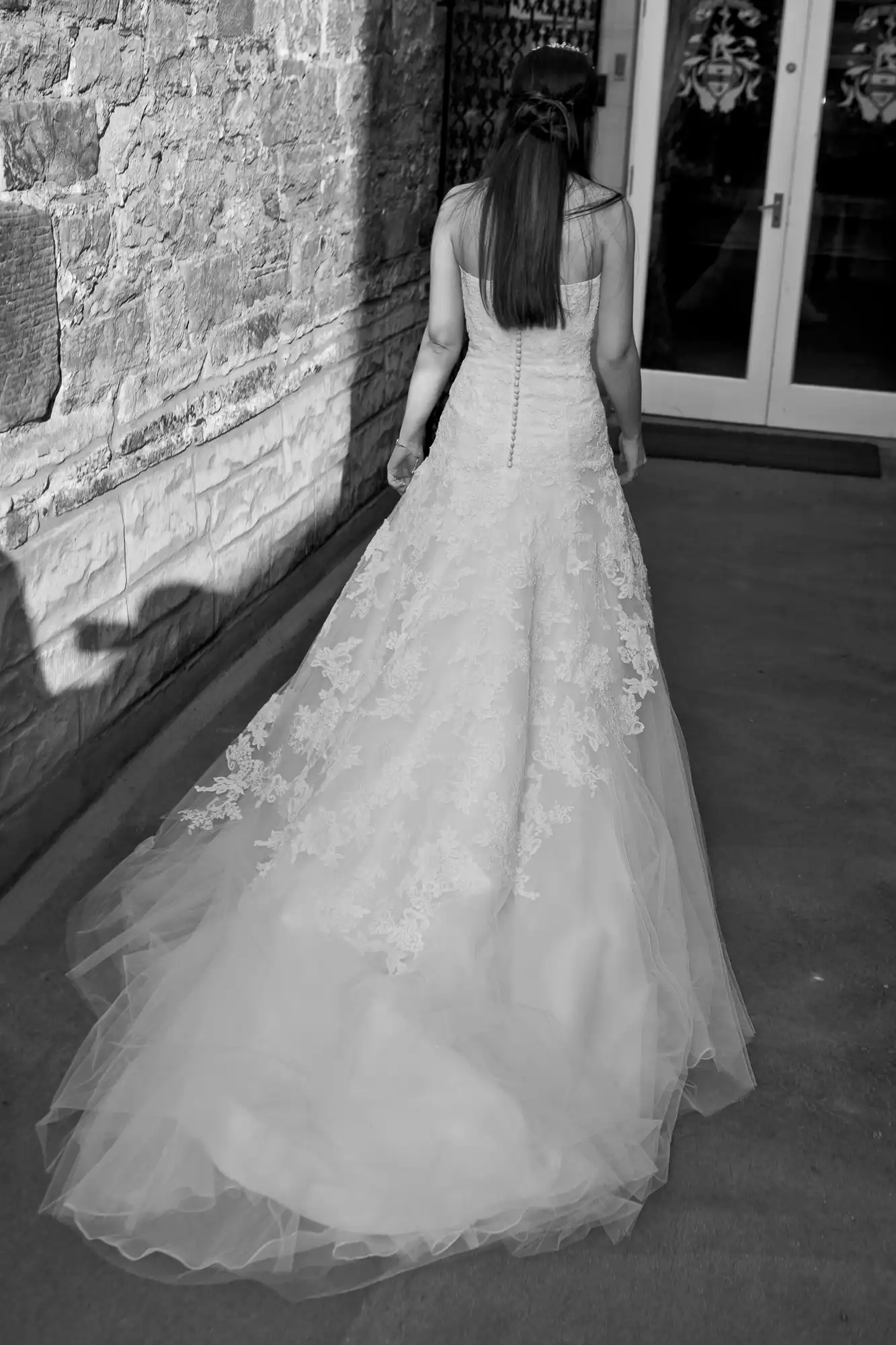 A woman in a lace wedding dress walking away from the camera, with a focus on the detailed gown and train, in black and white.