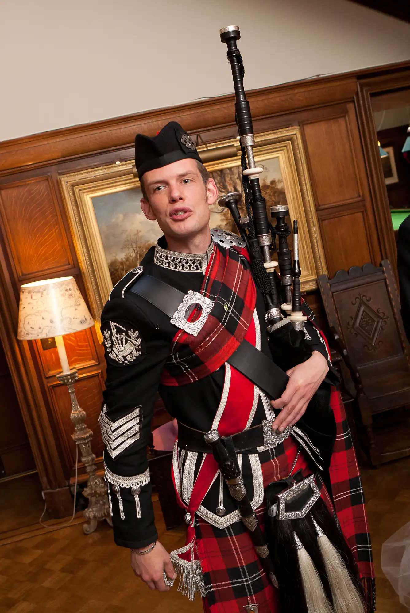 Man in traditional scottish attire, including a kilt and sporran, holding bagpipes, standing in a warmly lit room with vintage decor.