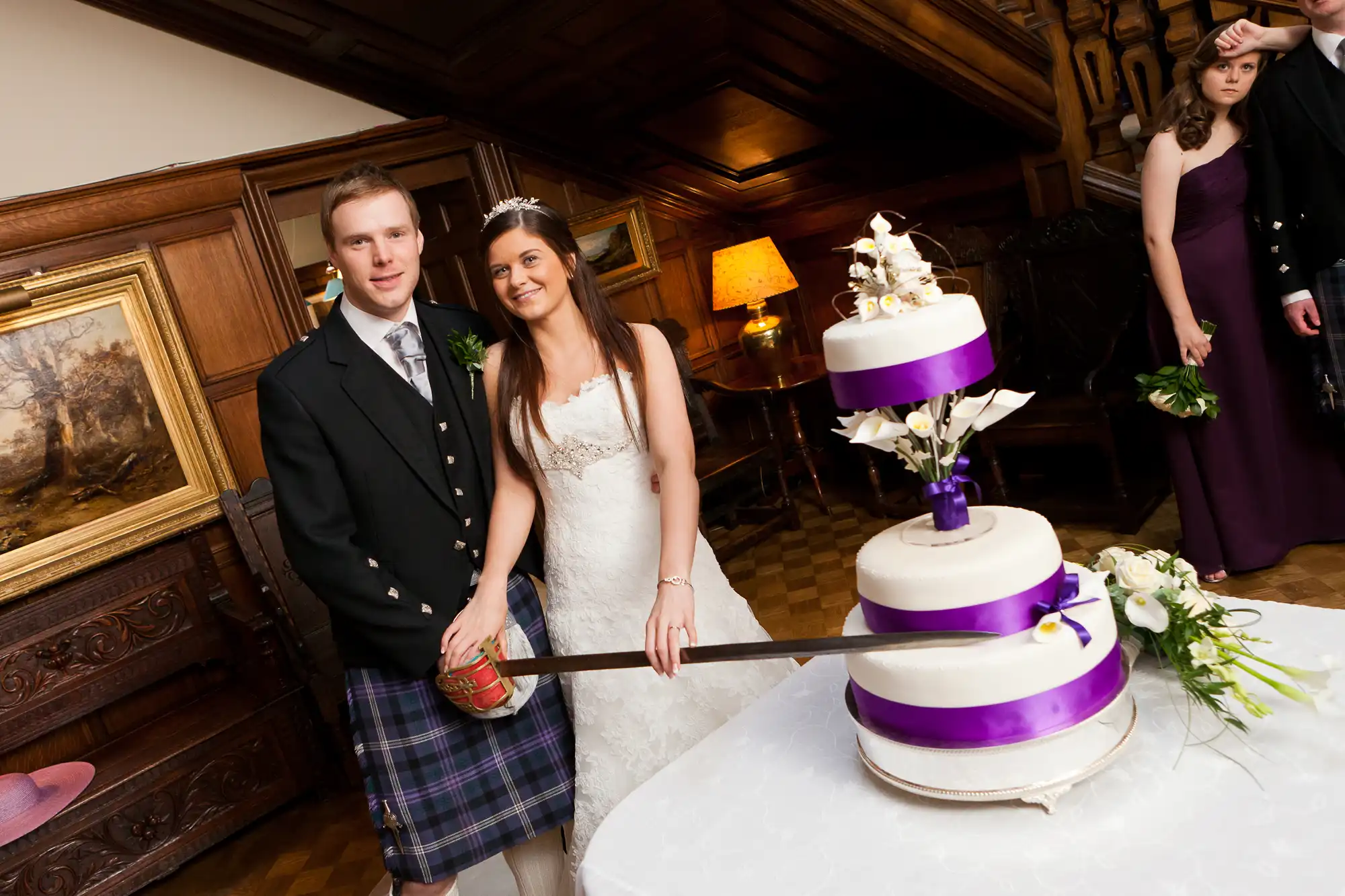 A bride and groom in formal attire cut a white and purple wedding cake in an elegantly decorated room.