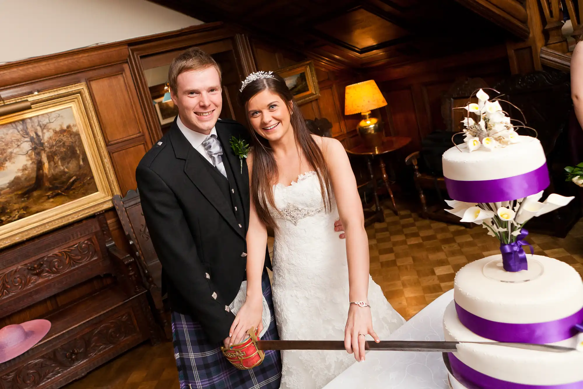 A bride and groom in wedding attire, smiling and cutting a white and purple tiered cake in a warmly lit room with wood paneling.
