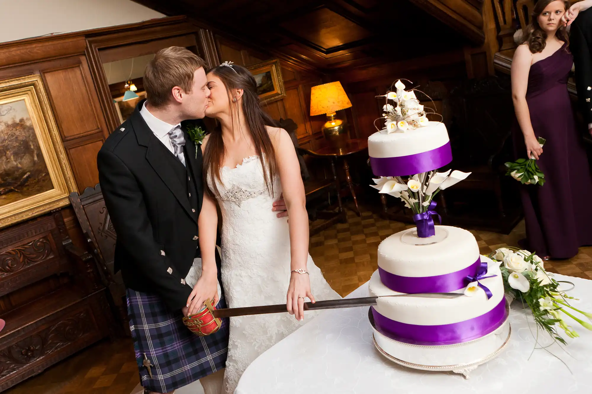 Bride and groom in wedding attire kissing beside a three-tiered white and purple cake in a warmly lit room, with a bridesmaid looking on.