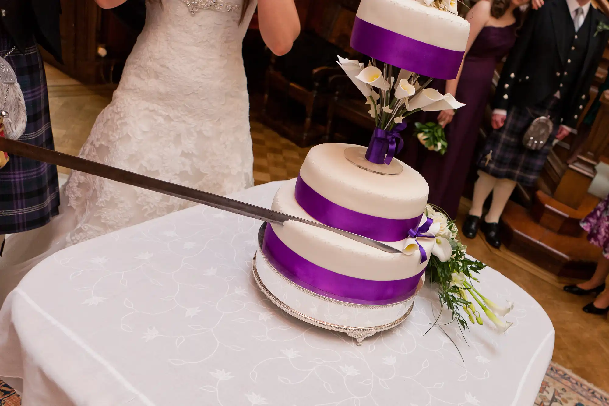 A newlywed couple cutting a three-tier wedding cake with purple ribbons and white floral decorations at a reception.