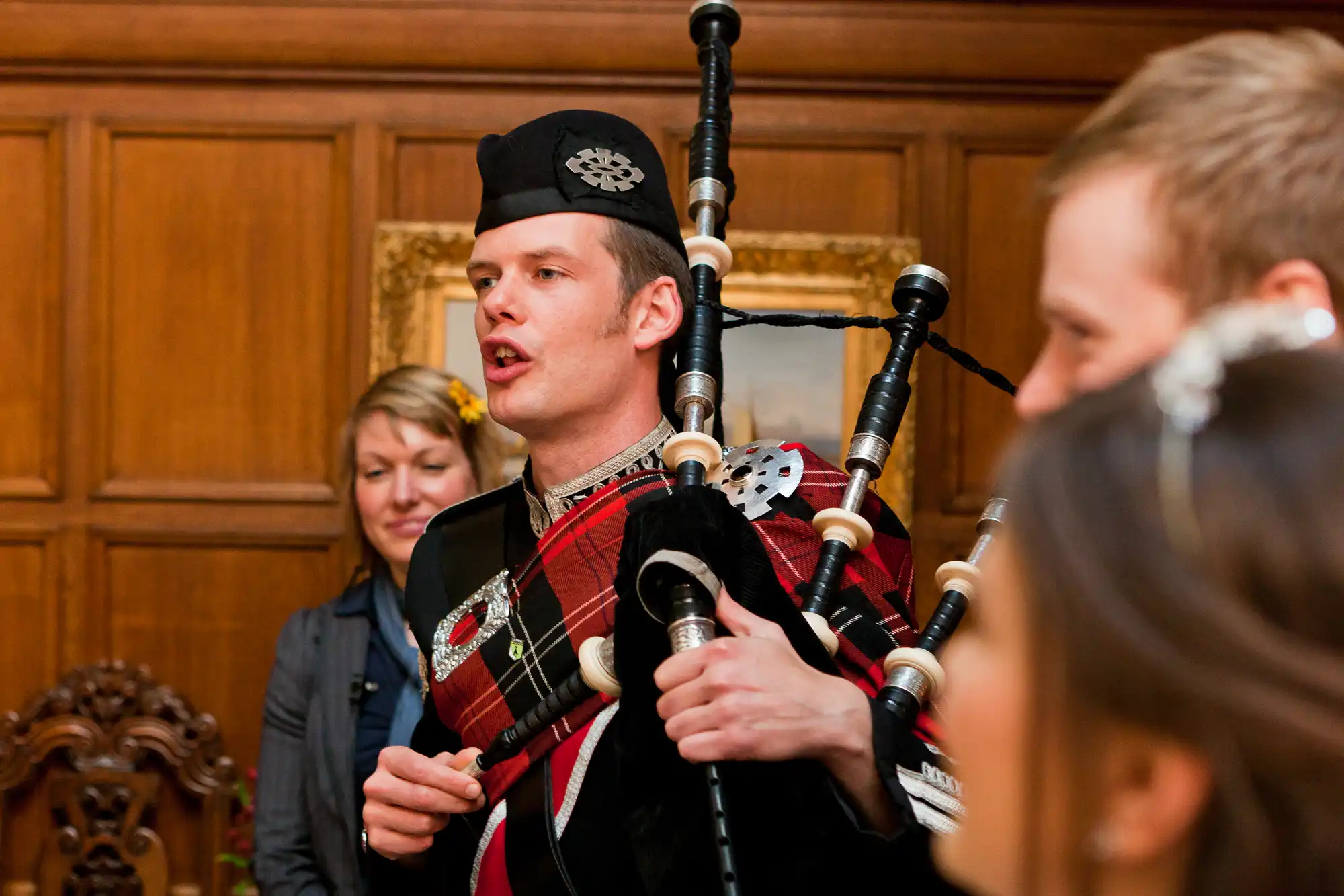 A man in traditional scottish attire playing the bagpipes at an indoor event, with two people observing in the background.
