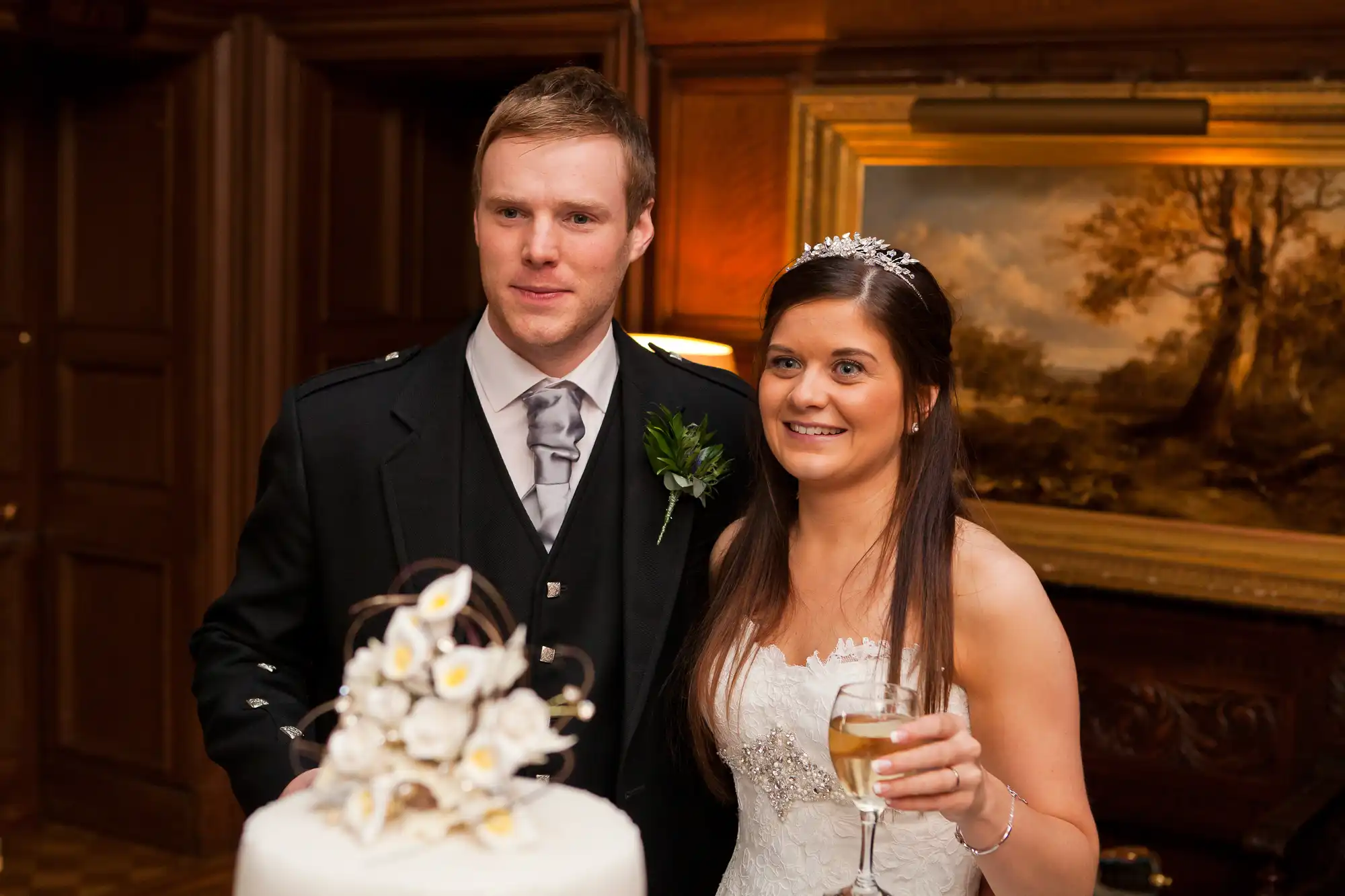 A bride and groom standing beside their wedding cake in an elegant room, smiling gently as they prepare to cut the cake.