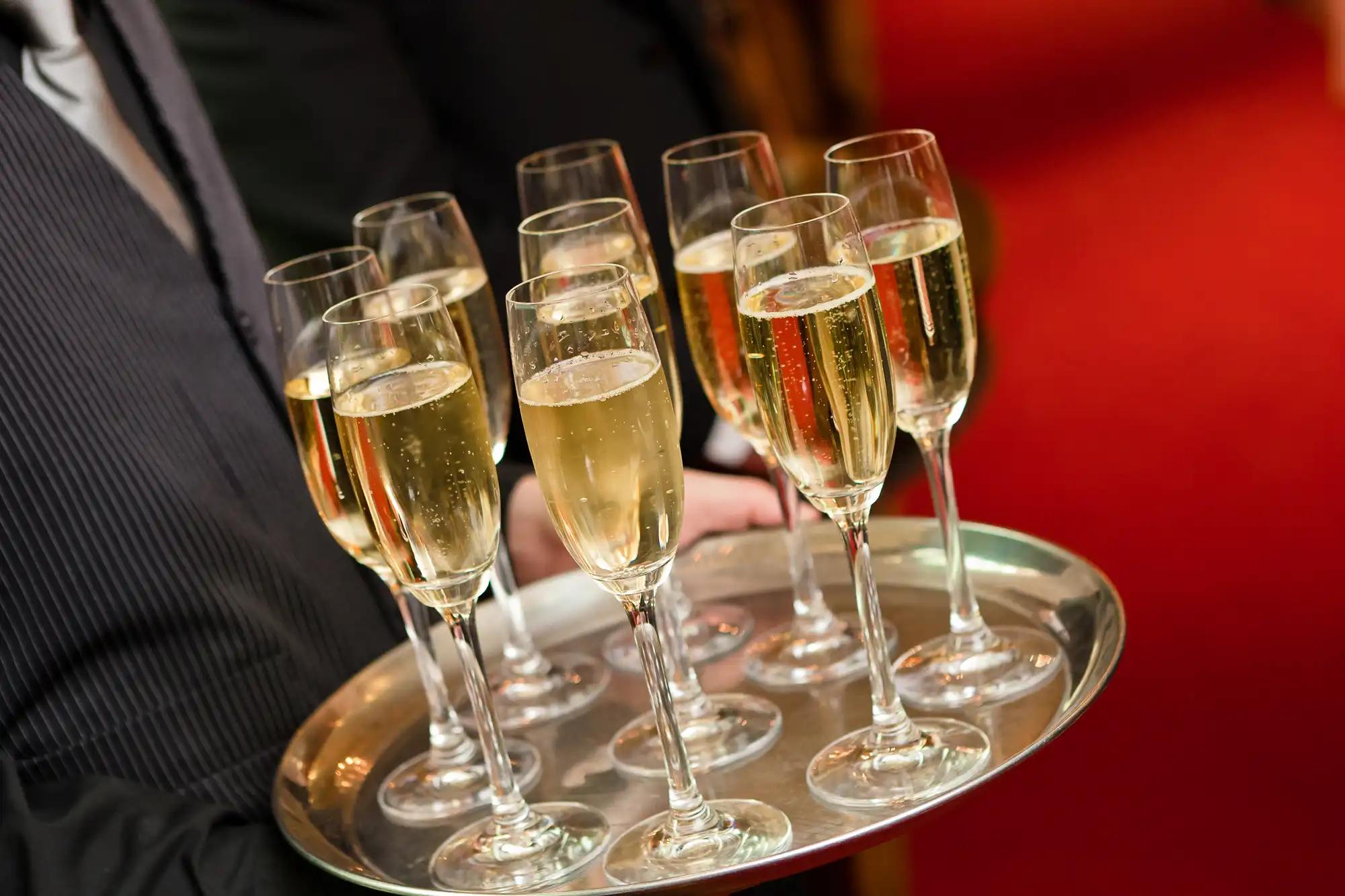 A server holding a tray of champagne glasses at a formal event with a red carpet background.