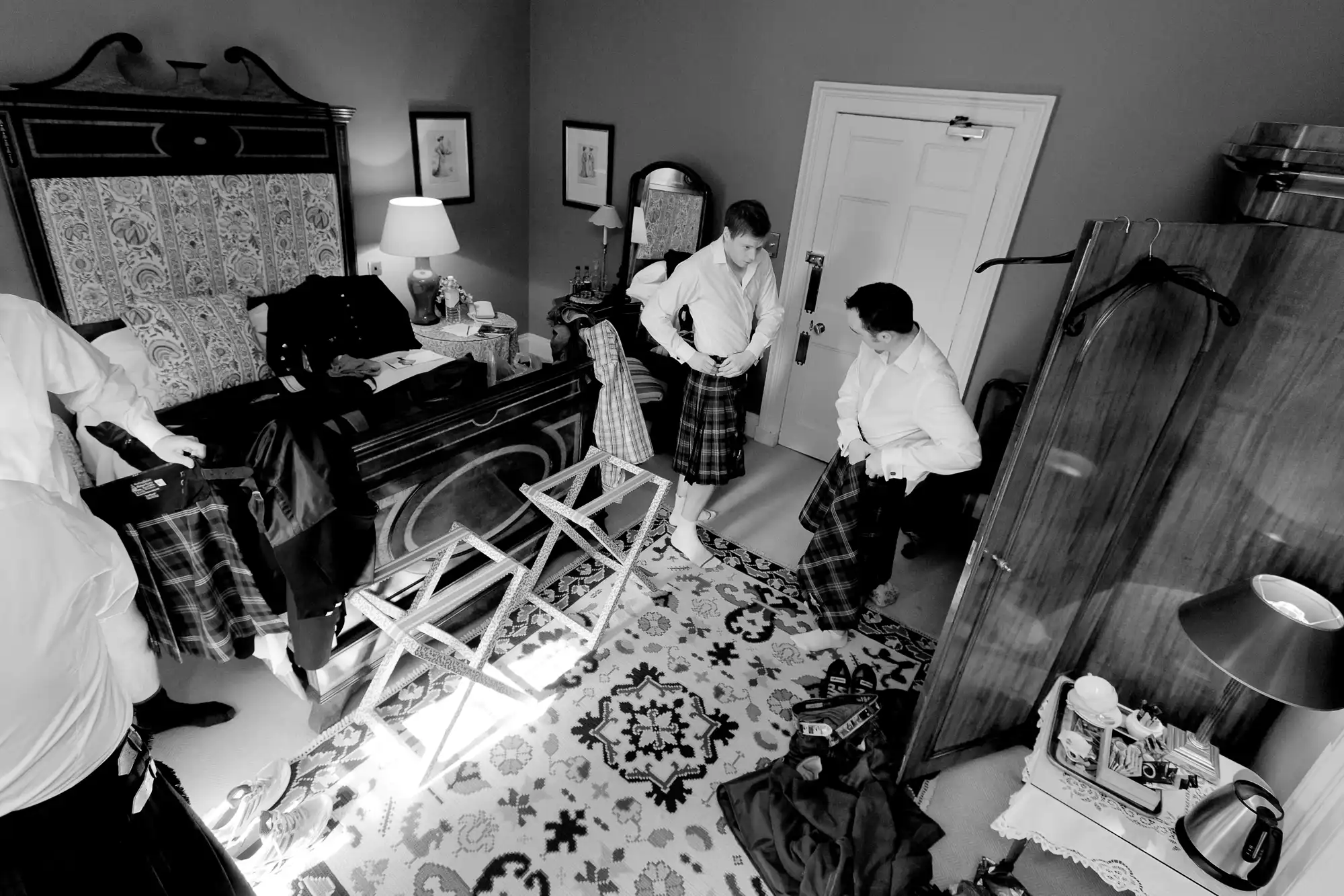 Two men in kilts preparing in a cluttered room, one adjusts the other's attire.