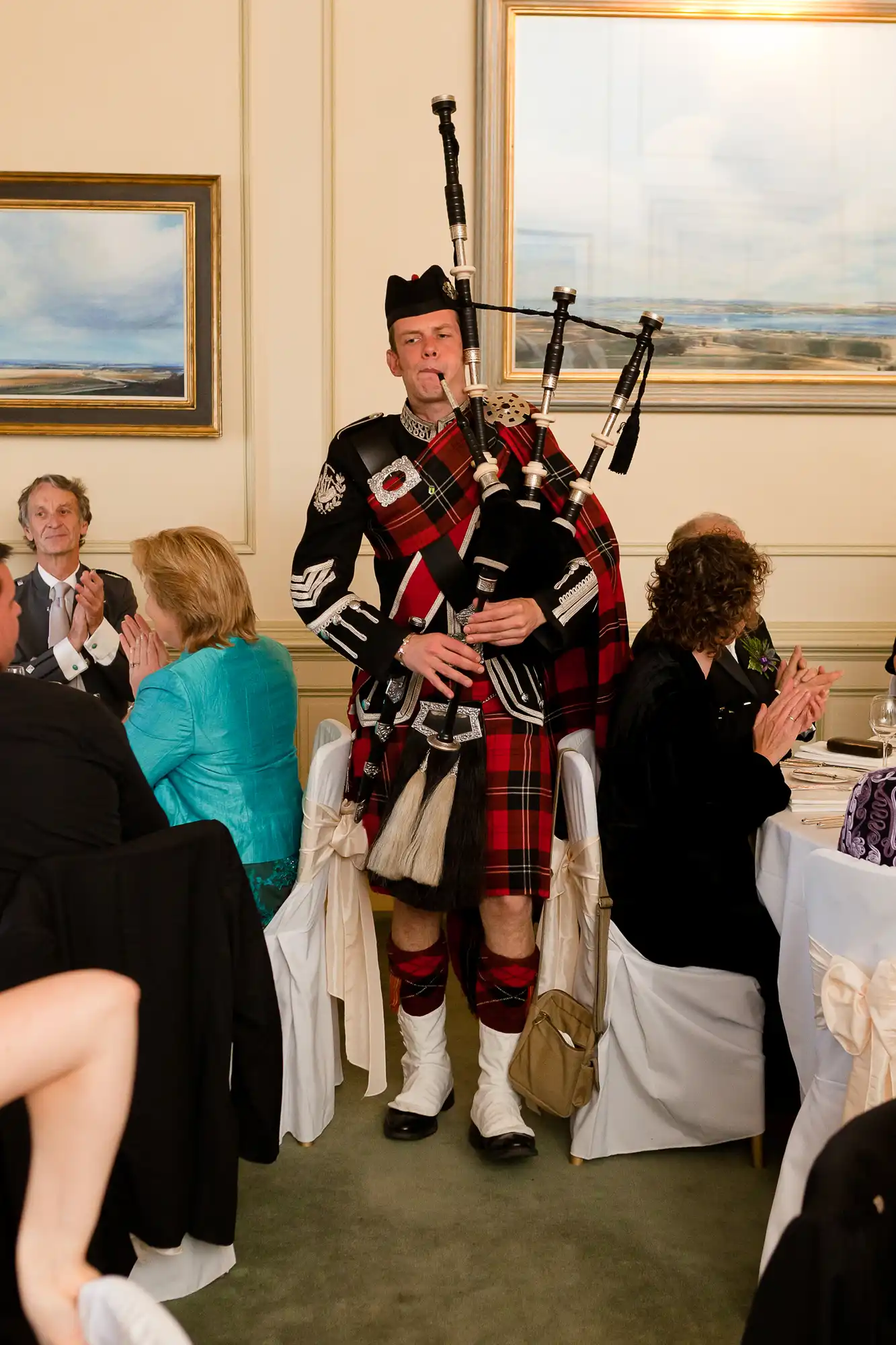 A bagpiper in traditional scottish attire playing bagpipes at an indoor event, surrounded by seated applauding guests.